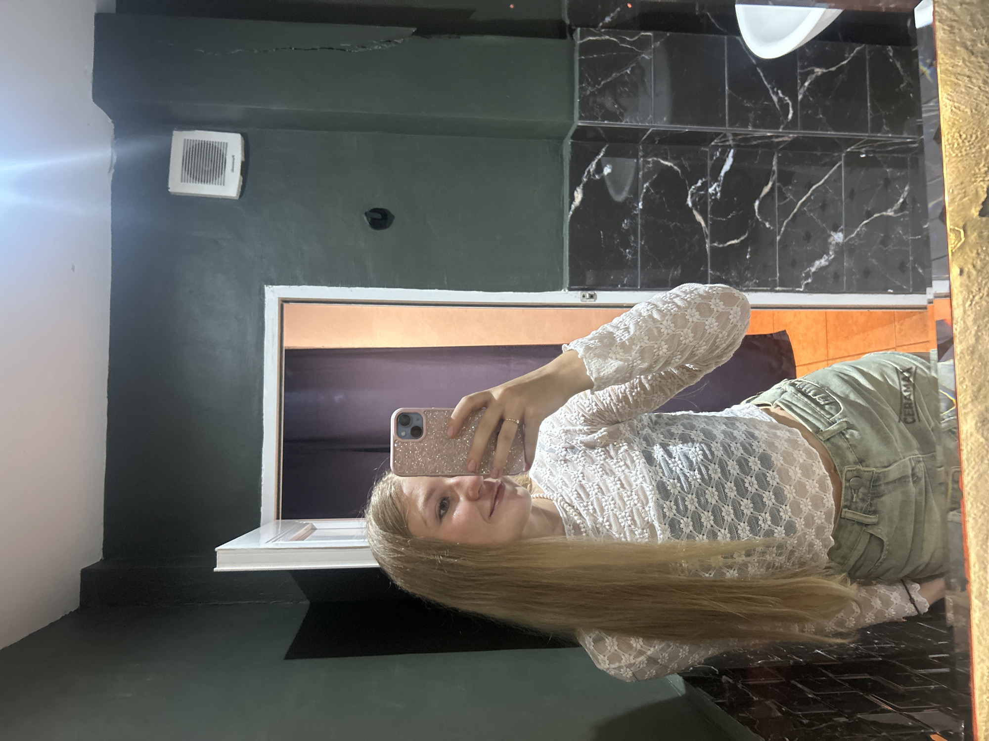 A young woman with long, blonde hair is taking a selfie in what appears to be a bathroom. She is holding a pink phone with a glittery case. She's wearing a white, lacy, long-sleeved top and light-colored jeans. The bathroom has dark green walls, some with black marble tile. There's a vent near the ceiling and a window behind her. The location is identified as Kuta Selatan, Indonesia.