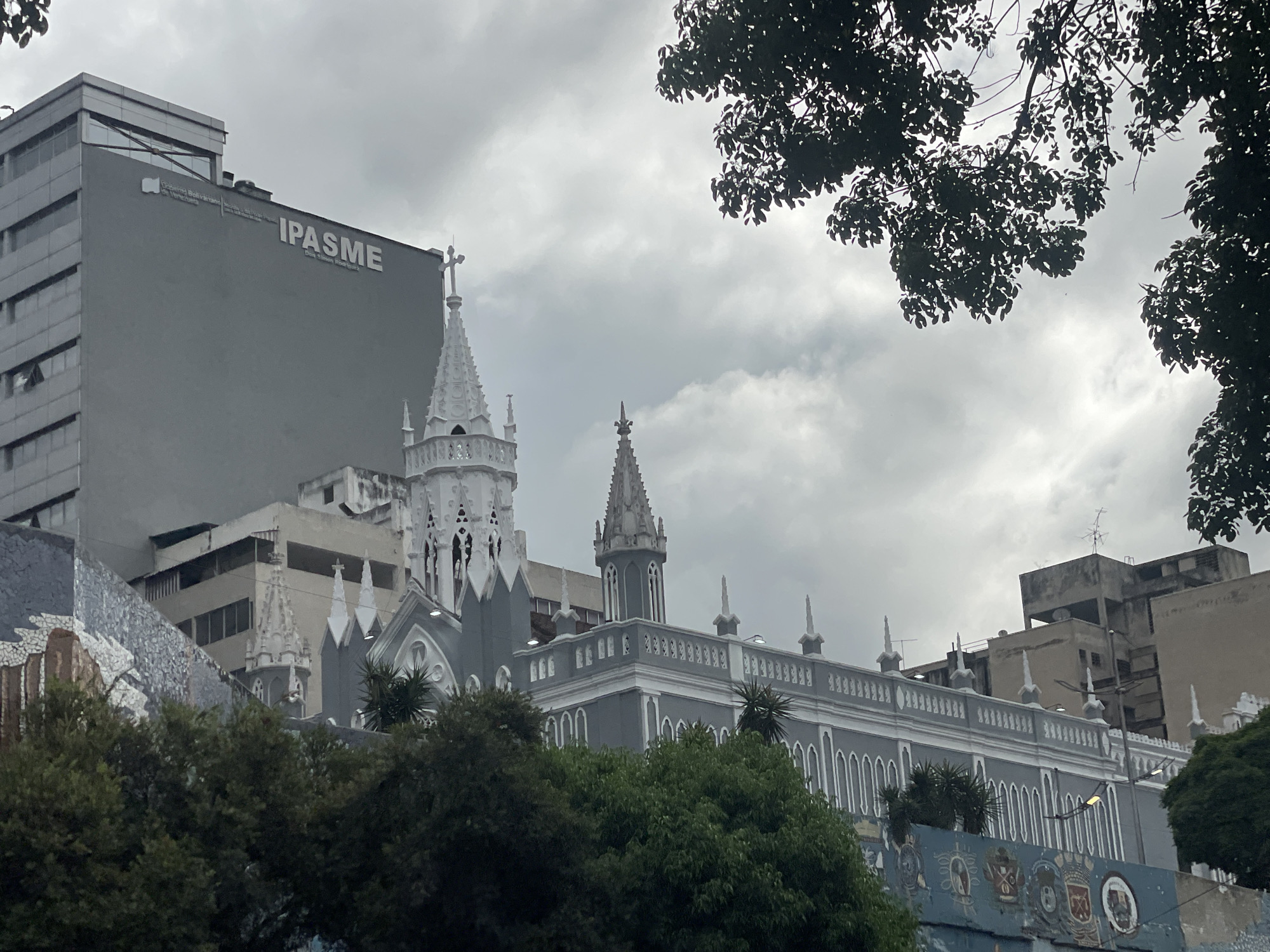 The image shows a view of Caracas, Venezuela, on a cloudy day. In the foreground, trees partially obscure a building with murals. Behind this, the grey and white neo-gothic style "Iglesia de San Francisco" is visible, with its spires reaching towards the sky. To the left of the church, a tall, grey building with the inscription "IPASME" is present. Other buildings are visible in the background. The overall scene conveys an urban environment with a mix of architectural styles.