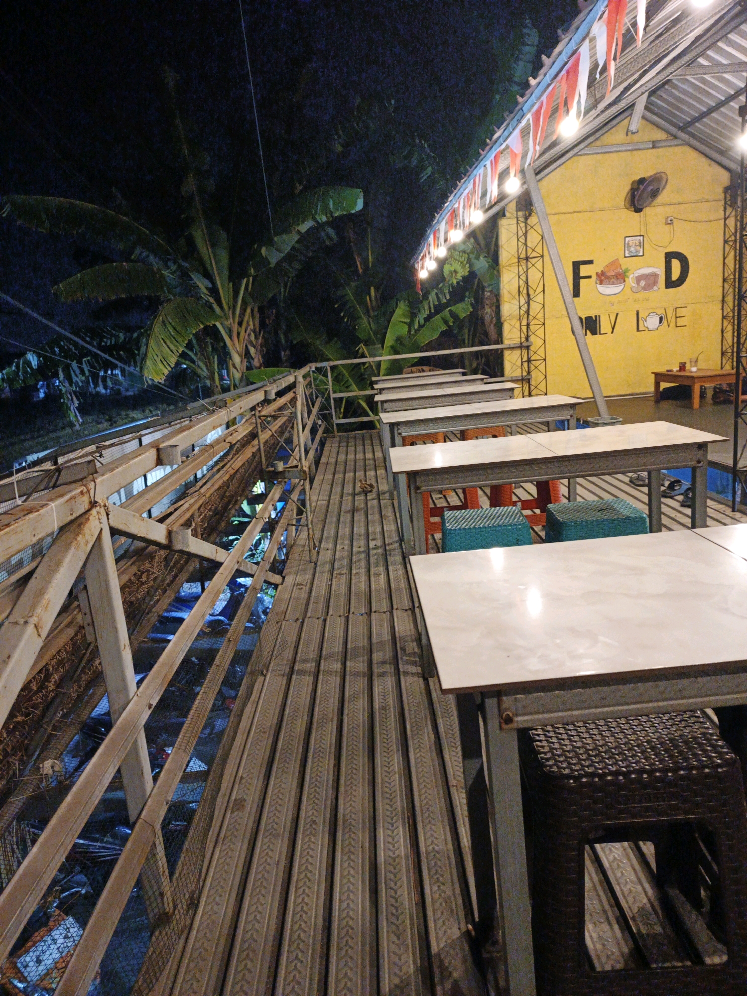 A nighttime outdoor scene shows a deck with tables and chairs at a restaurant called "FOOD ONLY LOVE" in Ketegan, Indonesia. The restaurant building is painted yellow and decorated with red and white Indonesian flags and string lights. Banana trees are visible behind the restaurant. The deck is made of metal and wooden planks, with metal railings on the left side. Several tables with white surfaces and metal legs are set up, with chairs of various colors. The scene appears to be empty.