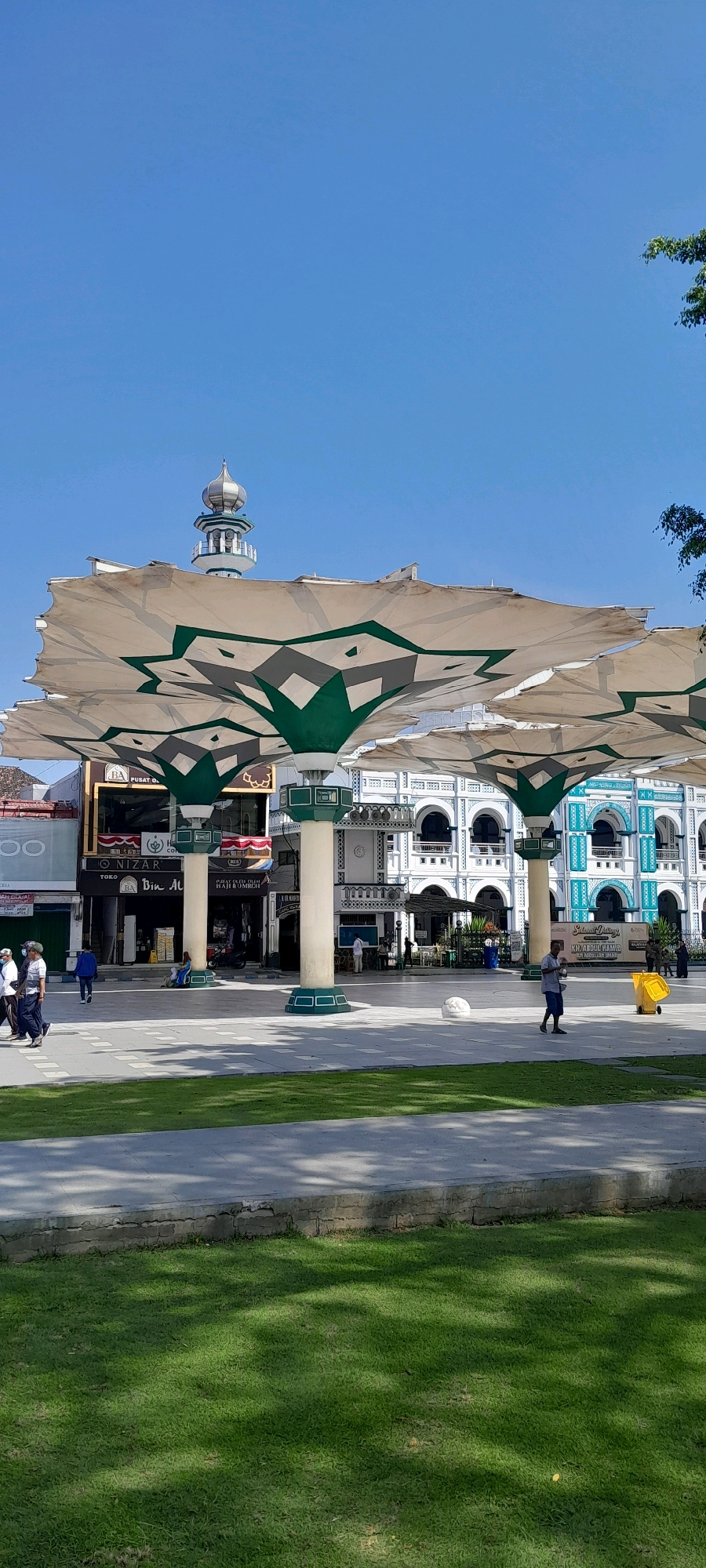The image captures a sunny day in Pasuruan, Indonesia, likely near a significant mosque or public square. Prominently featured are several large, retractable awnings with a distinctive star-like pattern in green and beige, supported by white pillars. In the background, a mosque with a dome and minaret is visible against a clear blue sky. Adjacent to the awnings are buildings with ornate facades, some painted white and others in a vibrant turquoise and white combination. The foreground shows a paved walkway and a well-maintained grassy area. Several individuals are present, including pedestrians walking and a person pushing a yellow bin. The scene suggests a public gathering space, possibly a religious or cultural site.