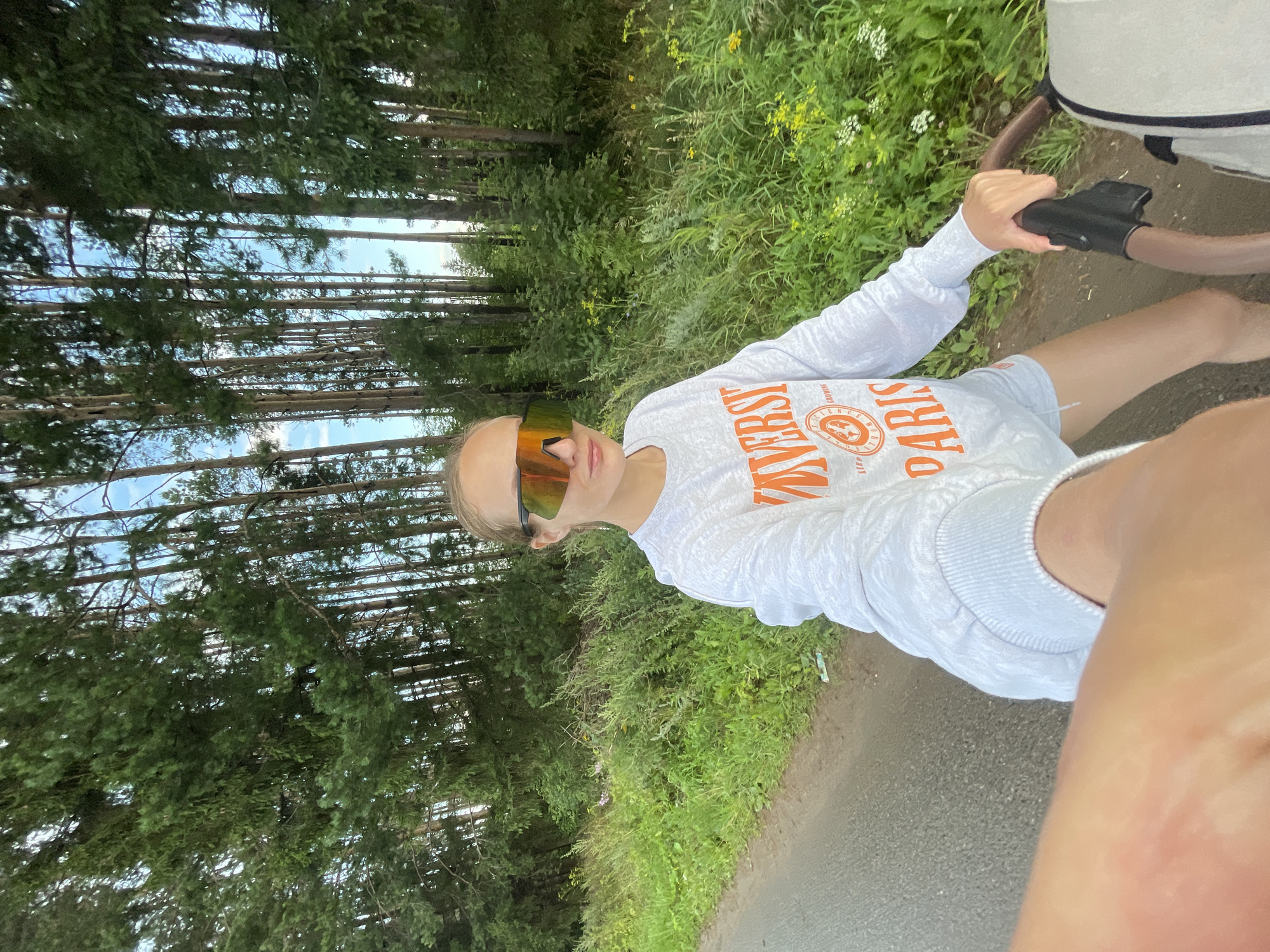 A woman with blonde hair pulled back and wearing large, mirrored sunglasses and a light gray sweatshirt with orange lettering is walking down a paved road. The sweatshirt has "UNIVERSITY" and "PARIS" printed on it, along with a circular logo containing an image of the Eiffel Tower. She is holding the handle of a stroller, the top portion of which is visible in the lower right corner of the frame. The road is bordered by a grassy verge with wildflowers. Behind the verge is a dense forest of tall, slender pine trees under a bright, partly cloudy sky. The scene appears to be located in or near the village of Podshivalovo, Russia, given the presence of pine trees and a dirt road.