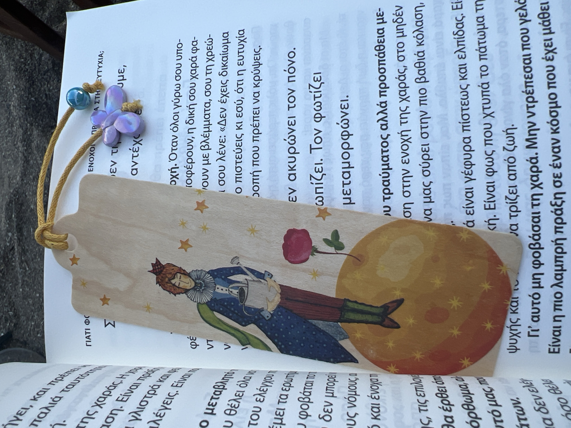 The image shows an open book with a bookmark placed inside. The bookmark is made of light-colored wood and features an illustration of a character resembling the Little Prince, holding a watering can, standing on a yellow planet with red accents. The bookmark is adorned with a yellow cord and a small blue bead, along with a lavender butterfly charm. The book's pages display Greek text. The lighting suggests an outdoor setting, possibly a park or a public space, with shadows indicating natural light. The environment appears to be a relaxed setting, indicated by the informal arrangement of the book and bookmark. There is no information to locate this scene in Eretria Municipal Unit, Greece.