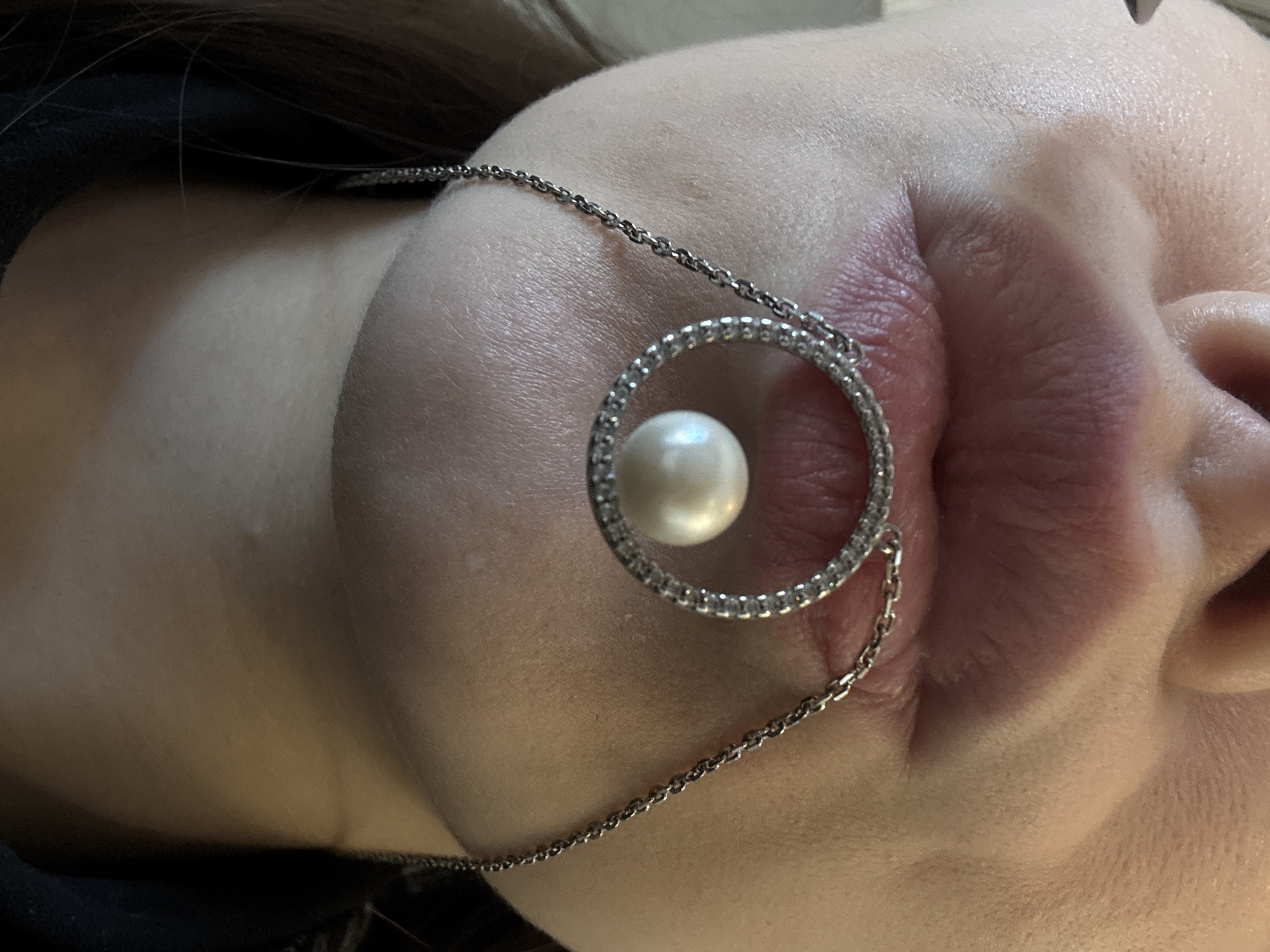 The image displays a close-up of a person's mouth and neck, adorned with a necklace. The necklace features a circular pendant with small, sparkling stones, and a single pearl suspended within the circle. The chain of the necklace is delicate and silver in color. The person's lips are slightly parted, and their skin appears fair. The background is a dark, indistinct color. No specific location within a city or country is discernible from the image.
