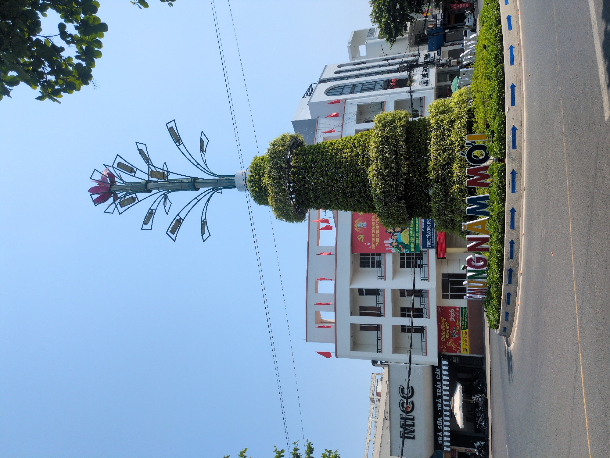 The image captures a street scene in Tuy Hòa, Vietnam. The main subject is a decorative structure with a central green topiary and a metal sculptural element resembling a stylized flower or plant. In front of the structure, there is a sign with the words "INGN MMO". The background consists of a building with red flags and banners, as well as other commercial establishments. A street with blue directional arrows is in the foreground. A tea shop named MICC is visible. Above, there's a clear blue sky with tree branches visible at the top corners of the image.