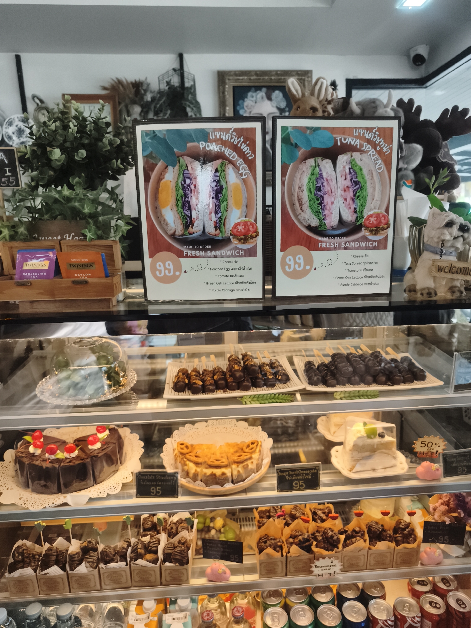 This image depicts the interior of a cafe, likely in Mai Khao, Thailand, showcasing a display case filled with various food items and promotional signage.
In the upper section, two framed menu boards are visible. The left board displays a picture of a sandwich with the text "Poached Egg" and the price "99.". The right board shows a similar sandwich with the text "Tuna Spread" and the price "99.". Both boards appear to be in Thai and English. Behind the menu boards, several stuffed animal toys are placed, including what looks like a rabbit, an elephant, and a moose. A small white dog figurine with a blue collar, labeled "welcome", is positioned to the right of the menu boards.
The display case itself contains multiple shelves filled with food. On the top shelf, to the left, a slice of cake under a glass dome is visible. To the right of this, on the middle shelf, are several rows of chocolate-covered items on sticks, resembling skewers of fruit or pastries, on white trays. Further to the right on this shelf, a slice of white cake with green and blue fruit on top is displayed under plastic wrap.
On the shelf below, a dark chocolate cake with red cherry toppings is on the left, with a price tag of "95". To its right, a sliced cheesecake with a pretzel-like topping is visible, also with a price tag. Next to the cheesecake, a pink decorative item is placed.
The bottom shelf of the display case is filled with numerous small boxes, each containing what appear to be chocolate-covered pastries or candies. Some of these boxes have small labels. To the right of these, more promotional items and desserts are visible.
At the very bottom of the image, several cans of soda and bottled beverages are lined up, suggesting a retail or self-service area for drinks. The overall environment appears clean and well-lit, characteristic of a food establishment.