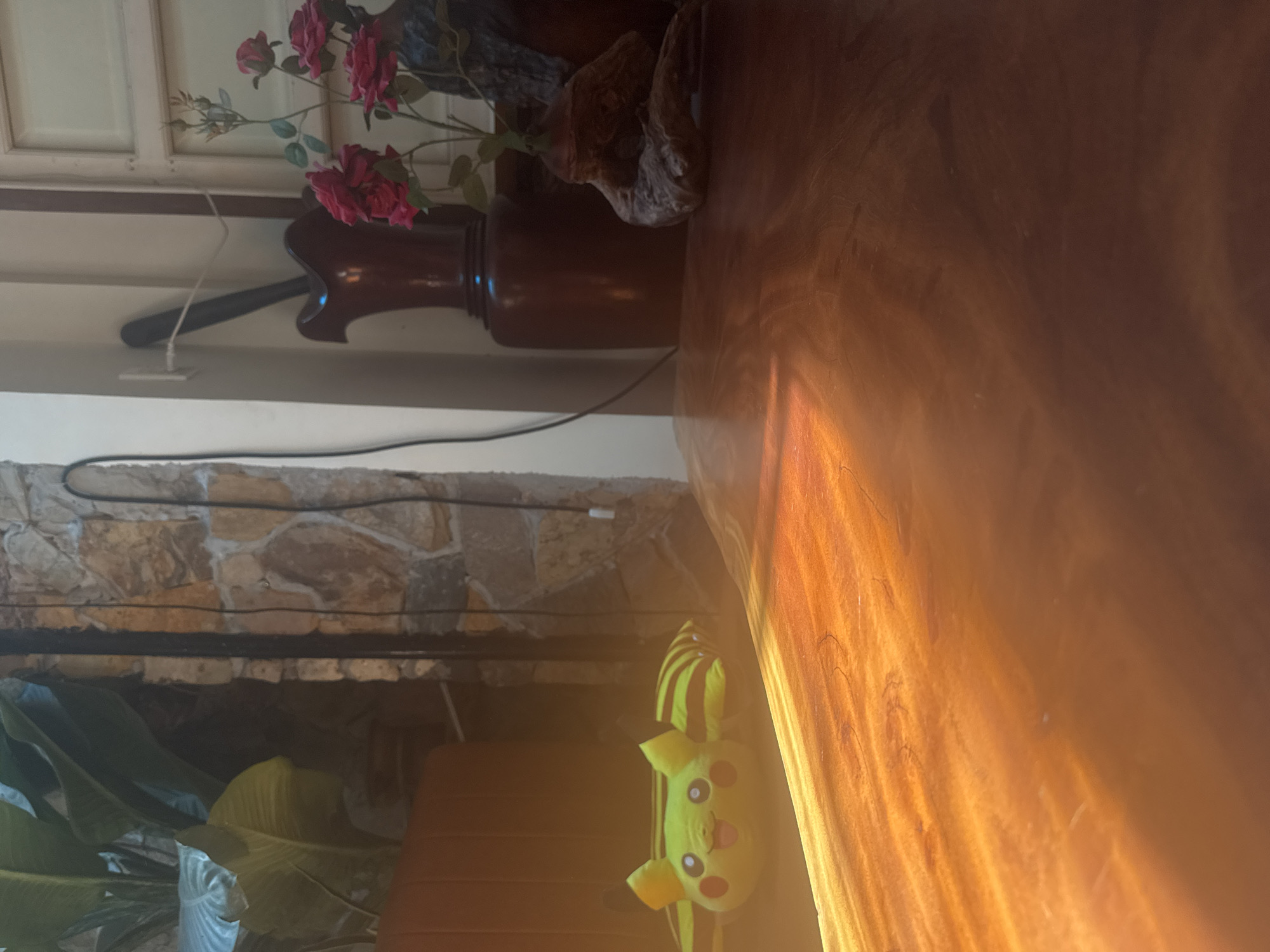 The image depicts an indoor scene with a focus on a wooden table illuminated by a shaft of sunlight. On the left side of the image, a Pikachu plush toy is visible, positioned near a brown couch or seating. Behind the plush toy and couch, a wall constructed of rough, multi-colored stones is apparent. A black cord runs vertically along the stone wall. To the right of the stone wall, a dark red vase stands next to a window with white panes. A few red roses in a decorative holder are also visible to the right of the vase. The foreground is dominated by the polished surface of the wooden table, reflecting the light in warm tones. The overall impression is of a domestic setting, possibly a living room or dining area. The city of Bac Nha Trang Ward, Vietnam, is mentioned as the location, but there are no specific visual cues in the image to confirm this geographical context.