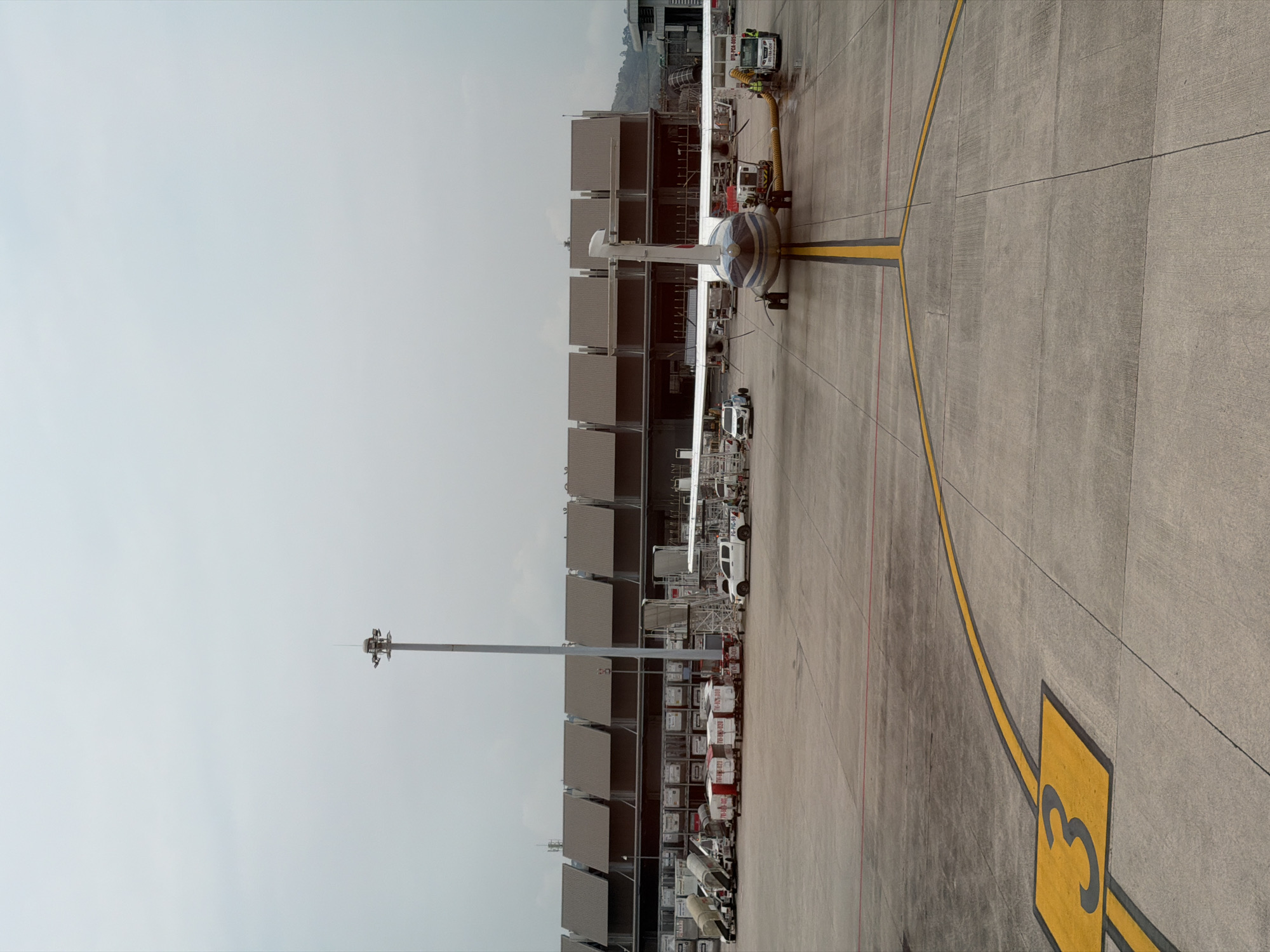 The image depicts an airport scene in Mai Khao, Thailand. A small passenger aircraft is parked at gate 3. The aircraft is white with blue striping on the nose and wings. Airport service vehicles are positioned around the aircraft, including a service truck connected to the aircraft with a hose. The aircraft is parked alongside a multi-story airport terminal building with numerous gates. The sky is overcast.