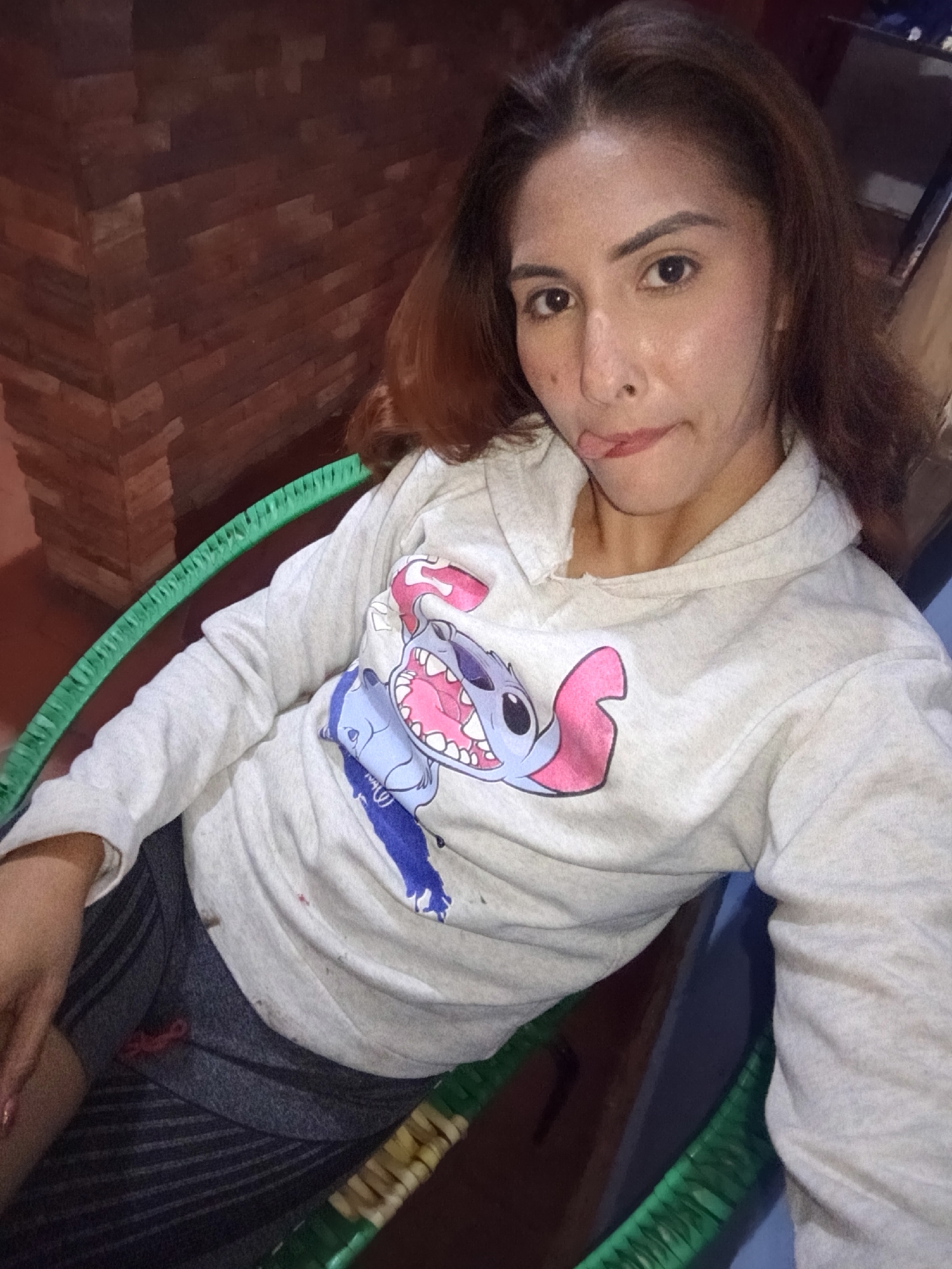 A young adult female is depicted in a medium shot, seated, taking a selfie. She has long, dark brown hair with lighter undertones, brown eyes, and fair to medium skin tone with visible pores and minor blemishes, including a notable mark below her left eye (viewer's left). Her expression is playful, with the tip of her tongue protruding between her slightly pursed, reddish lips. She is wearing a light gray, long-sleeved hooded sweatshirt with a graphic print featuring the blue cartoon character Stitch, shown with a wide-open mouth, prominent teeth, and pink inner ears, along with a smaller, partially visible blue character below it. She also wears dark gray shorts with subtle horizontal stripes. The female is seated in a chair with a distinctive bright green, woven or textured frame, and a woven light brown/beige seat. In the background to her left is a wall composed of reddish-brown, irregularly shaped rectangular elements resembling bricks or stone cladding. The overall lighting is moderate, suggesting an indoor or sheltered outdoor environment. The location is specified as Juan Griego, Venezuela.
