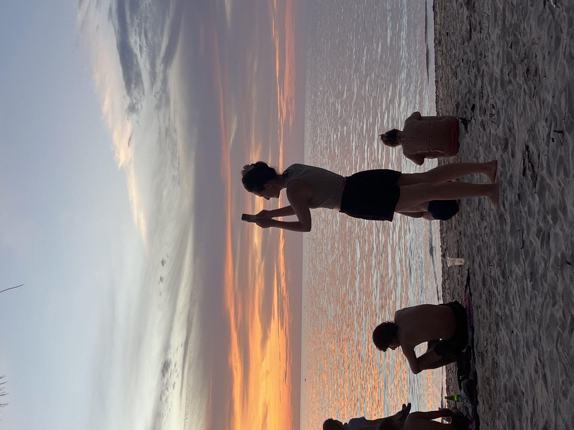 A group of people are silhouetted on a sandy beach at dusk. The sky is filled with vibrant orange and pink clouds, reflecting on the calm water of the sea. A woman in the foreground is standing and taking a photo with her phone, facing the sunset. Other individuals are seated on the sand, facing the water or looking away. The scene appears to be in Ko Pha-ngan District, Thailand.
