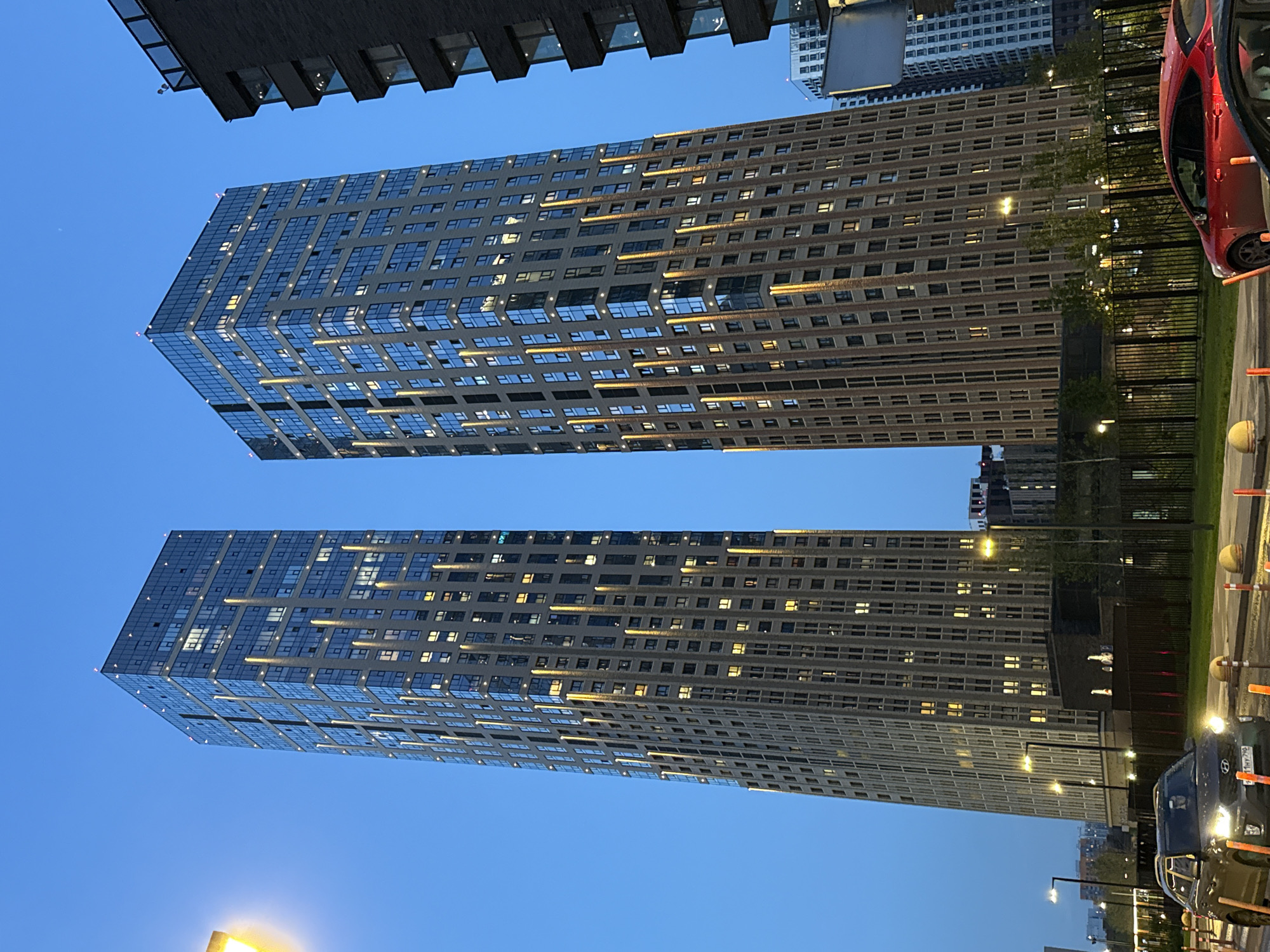 The image shows two tall, modern buildings in Khimki, Russia, illuminated by interior lights and reflecting the blue evening sky. The buildings feature a grid-like facade with numerous windows. Lower buildings are also visible in the background, and a few trees line a small patch of grass. In the foreground, there are a few cars, street lights, and traffic cones. The perspective is looking upwards, emphasizing the height of the buildings.