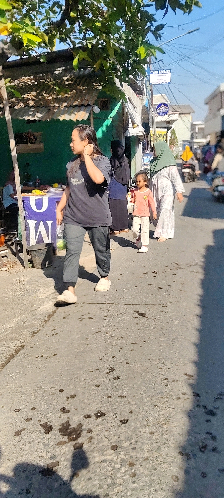 A medium-shot, vertical, outdoor photograph depicts a street scene under bright sunlight, likely in Mekarmukti, Indonesia. In the foreground, a woman with dark hair, wearing a dark grey t-shirt and dark grey pants, is walking to the left and looking towards the right. She is wearing off-white slip-on shoes. Her right hand is touching the back of her head. To her right, a young child, possibly a girl, in a light orange long-sleeved shirt and patterned light grey pants, is walking to the right. She is wearing light pink shoes. Behind the child, a woman wearing a light grey and white patterned dress and a light green hijab is walking to the right. Further back, a woman in a dark blue dress and black hijab is walking to the right. On the left side of the image, a wooden market stall with a purple front is visible. The stall has the letters "W" and "M" on its front. A dark grey bucket is placed next to the stall. In the background, there are buildings, a power line, and various signs, including one that reads "ATM BERSAMA". The road surface is asphalt and appears somewhat rough, with scattered dark spots. The overall scene suggests a typical street in a residential or commercial area.