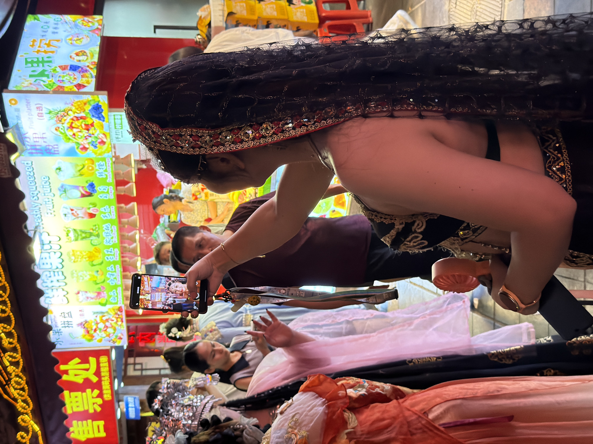 In this image, a woman in a black headscarf with red and gold trim is leaning forward, taking a photo with her phone. Her arm is extended, and the phone screen shows a picture of a woman and a man smiling. She is wearing a dark dress with gold embellishments. Behind her, a market scene unfolds. Brightly colored signs with Chinese text and images of fruit platters and juices are visible, suggesting a food or beverage vendor. Several other people are present, some looking at the woman or the displays. One man in the background appears to be looking at the woman's phone. The setting seems to be an outdoor market or street, possibly at night given the artificial lighting from the signs. The overall mood is lively and bustling, characteristic of a busy marketplace. The visible text includes "售票处" (Ticket office) and "Fruit juice" (possibly with Chinese translations), and other Chinese characters that appear to be related to menu items or branding. The woman's posture and the act of taking a photo suggest she is capturing a memory or sharing her experience.