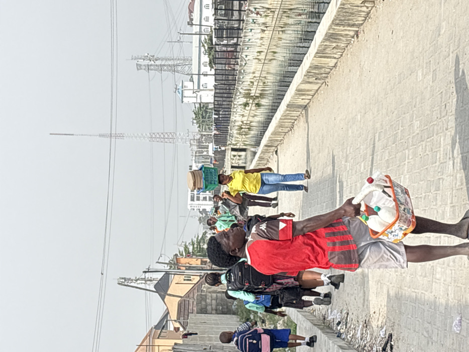 The scene depicts a paved pedestrian pathway in Igboefon, Nigeria, running parallel to a concrete-lined canal under a hazy, bright sky.
In the immediate foreground, a male individual, observed from a slight rear-left angle, wears a red and brown striped short-sleeved shirt, light grey shorts, and dark flip-flops. He holds a multicolored plastic basket containing multiple white plastic bottles with various colored caps (green, red, white) in his left hand. His head is turned slightly to the right, facing forward.
Behind this individual and to the left, a group of approximately five people, including several children with backpacks, walk away from the camera. One child on the far left wears a blue shirt and blue shorts with a blue backpack. Further down the pathway, a woman in a yellow short-sleeved shirt and blue jeans walks towards the camera, balancing a green basket containing light brown, circular objects on her head. Other indistinct figures are visible further along the path.
To the right, the canal contains dark, still water. The far bank of the canal features a dark fence line and vehicles on an unseen road, backed by multi-story buildings, including a white structure with a blue roof. To the left of the pathway, a concrete wall separates it from residential-style buildings with varied roof colors, and debris is visible at the base of this wall.
The background is dominated by a hazy, light grey sky. Multiple utility poles with overhead power lines crisscross the scene. Several prominent metal lattice towers, consistent with telecommunication or transmission structures, extend into the sky, with one slender tower on the left and a larger, broader structure further right.