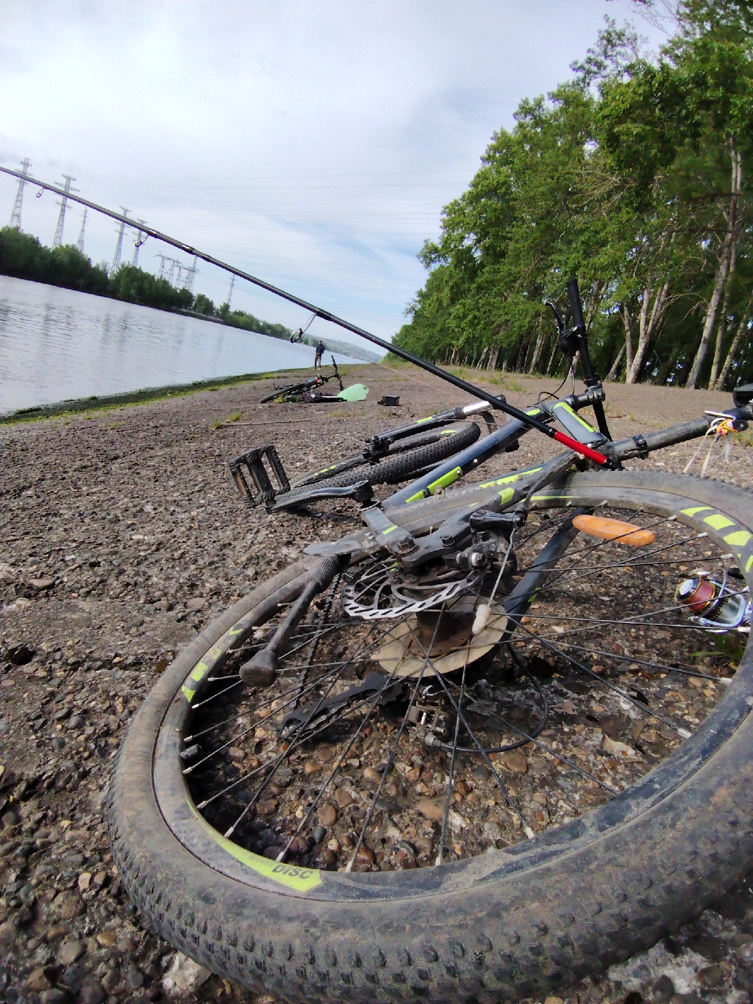 The image depicts a riverside scene in what appears to be Chaikovsky, Russia, indicated by the presence of power lines crossing a riverbank. Two bicycles are lying on a pebbly shore, one in the foreground and another further away. The bike in the foreground is prominently displayed, showing a disc brake, tire, and a fishing reel attached to the frame. Two fishing rods are positioned to extend over the water, suggesting someone is engaged in fishing. A person can be seen standing further down the shore. The sky is overcast. The river has still water. There is a row of green trees on the side of the river.