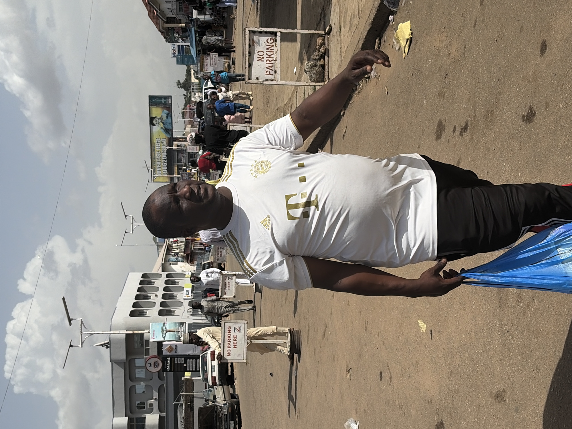 The image shows a man in a white and gold Adidas t-shirt and black shorts walking on a dusty street in Jos, Nigeria. He is carrying a blue plastic bag. In the background, there are buildings, cars, and other pedestrians. There are also signs that say "NO PARKING HERE". The sky is cloudy. The scene appears to be a busy marketplace or commercial area.