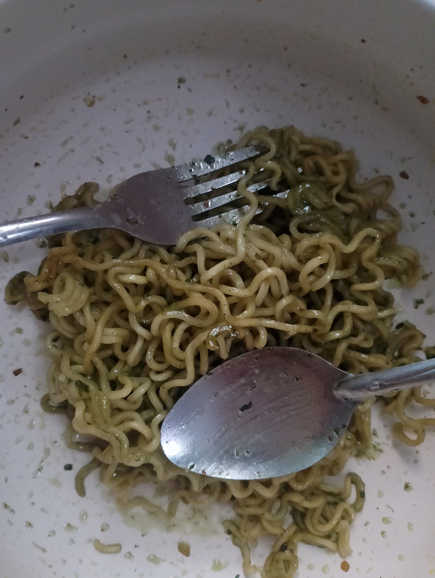 A close-up, overhead shot captures a portion of instant noodles in a bowl. The noodles, appearing slightly oily and green from seasoning, are piled in the center. A fork rests on the upper left portion of the noodles, its tines embedded slightly. A spoon is positioned in the lower right, partially covering the noodles. The bowl's white surface is speckled with remnants of the food. The image is taken in Magelang, Indonesia.