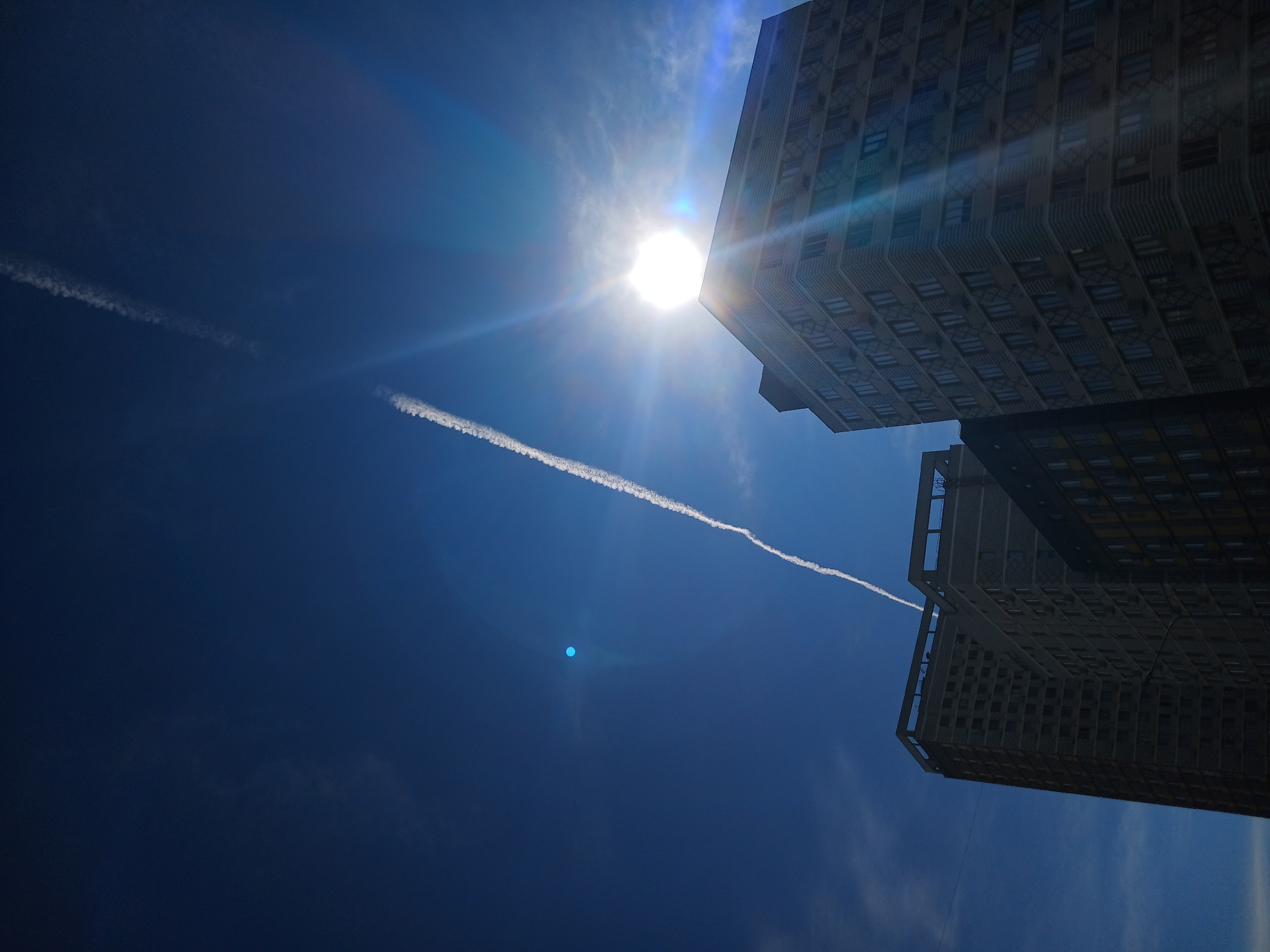 The image captures a perspective looking up at two multi-story buildings against a clear, deep blue sky. A bright sun is prominently positioned in the upper right quadrant of the frame, emitting strong lens flares and light rays that cast over the facade of the building on the right. A white contrail from an aircraft stretches diagonally across the sky from the upper left to the lower right. The buildings appear to be residential or office buildings, with visible windows and some architectural details. The building on the left is partially under construction or unfinished, with a more bare exterior compared to the finished building on the right. The scene is set in the city of Yekaterinburg, Russia, indicated by the context provided. There are no people or overt actions depicted in the image; it is a still shot of architecture and sky.