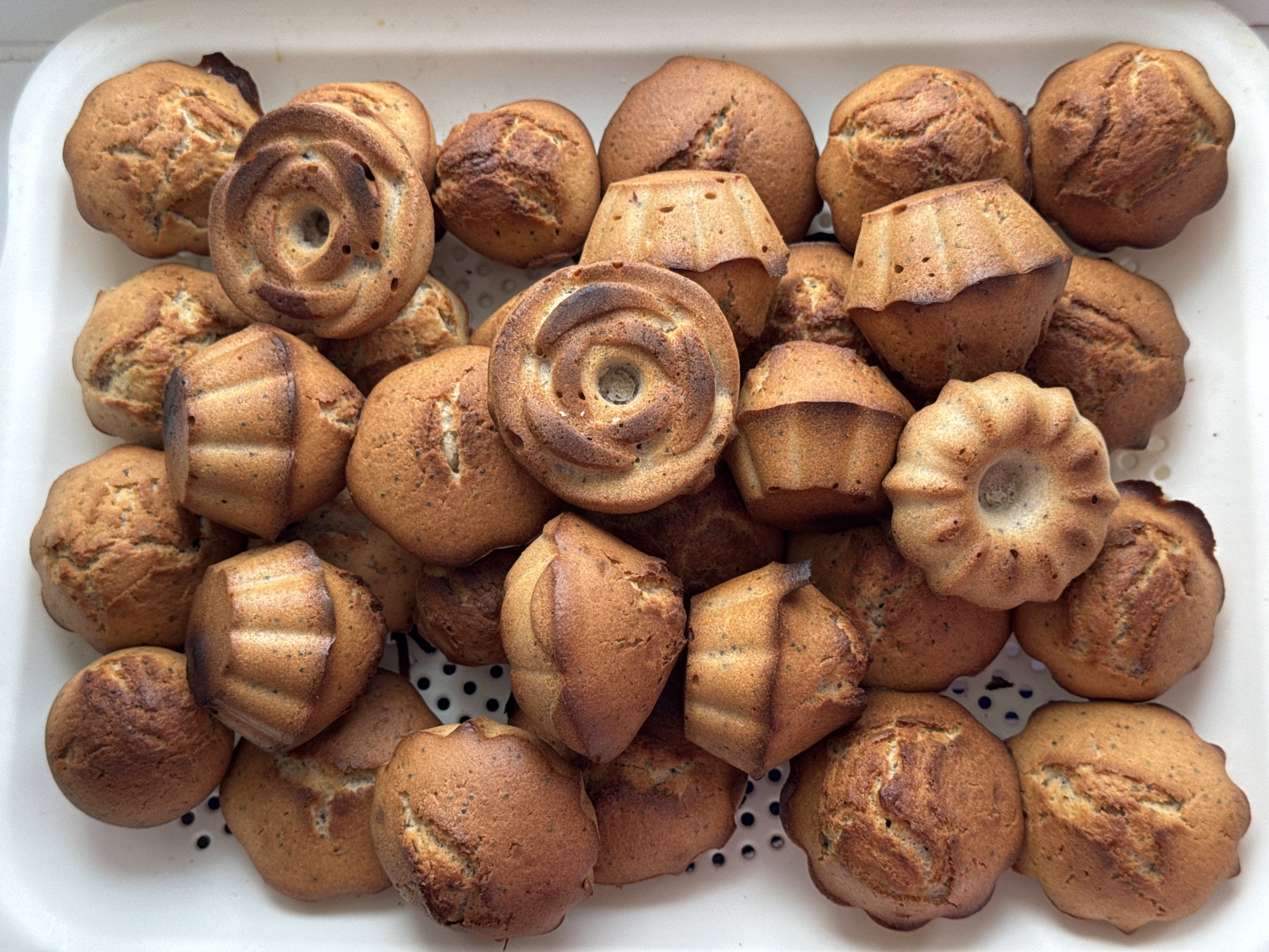A high-angle shot shows a white tray filled with an assortment of muffins. The muffins are of different shapes, some round, some with flower-like designs, and some with a distinctive geometric shape. They are uniformly tan in color with subtle variations in shading. The texture of the muffins appears slightly cracked in places. The tray is white and has holes on the sides. The scene appears to be lit by natural light. The location is Высокий, Russia.