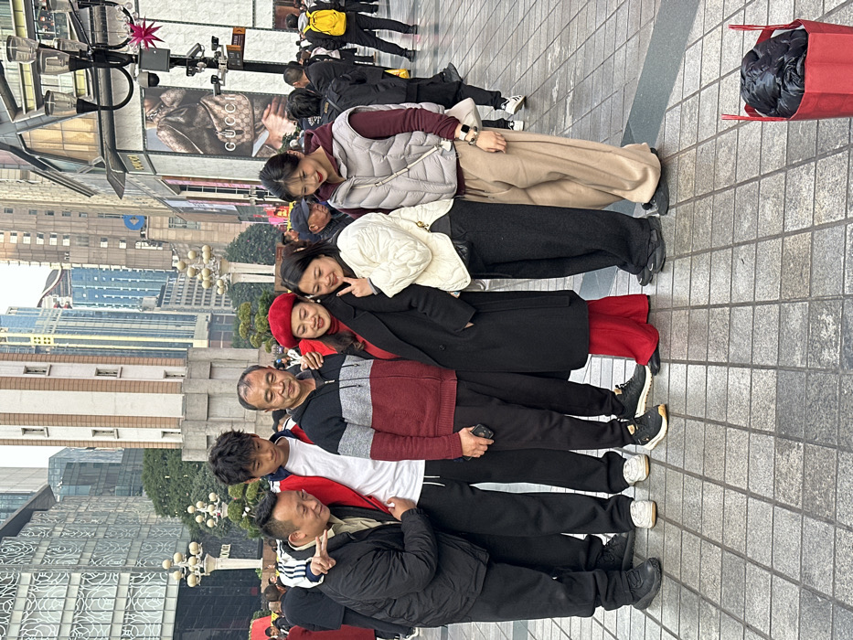 The image depicts six individuals, appearing to be of East Asian descent, posing for a photograph on a paved pedestrian street in the Jiefangbei commercial district of Yuzhong District, Chongqing, China. From left to right, the group consists of two adult men, one young male, and three adult women.
The leftmost man, wearing a black jacket, places his left arm around the shoulder of the young male, who is dressed in a white t-shirt with a red and white athletic jacket draped over his shoulders. The second man, wearing a grey, red, and black zip-up sweater, holds a smartphone in his right hand and has his left arm around the shoulder of the first woman. This woman is dressed in a black long coat and a red beret, and holds her right hand up in a "peace" gesture. To her right, the second woman wears a white quilted jacket and makes a "peace" gesture with her left hand. The third woman, to the far right, is dressed in a grey puffer vest over a maroon hoodie. All six individuals are either smiling or looking down subtly.
The background features several modern high-rise buildings under an overcast sky, including the distinctive Jiefangbei Monument. A large "GUCCI" store sign with a handbag advertisement is prominently displayed on a building to the right. Street lamps with multiple light fixtures and a red star decoration are visible. The ground is paved with square grey tiles. In the lower right corner, a red fabric bag contains a dark, folded item. Numerous other pedestrians are visible moving through the busy urban environment in the background.