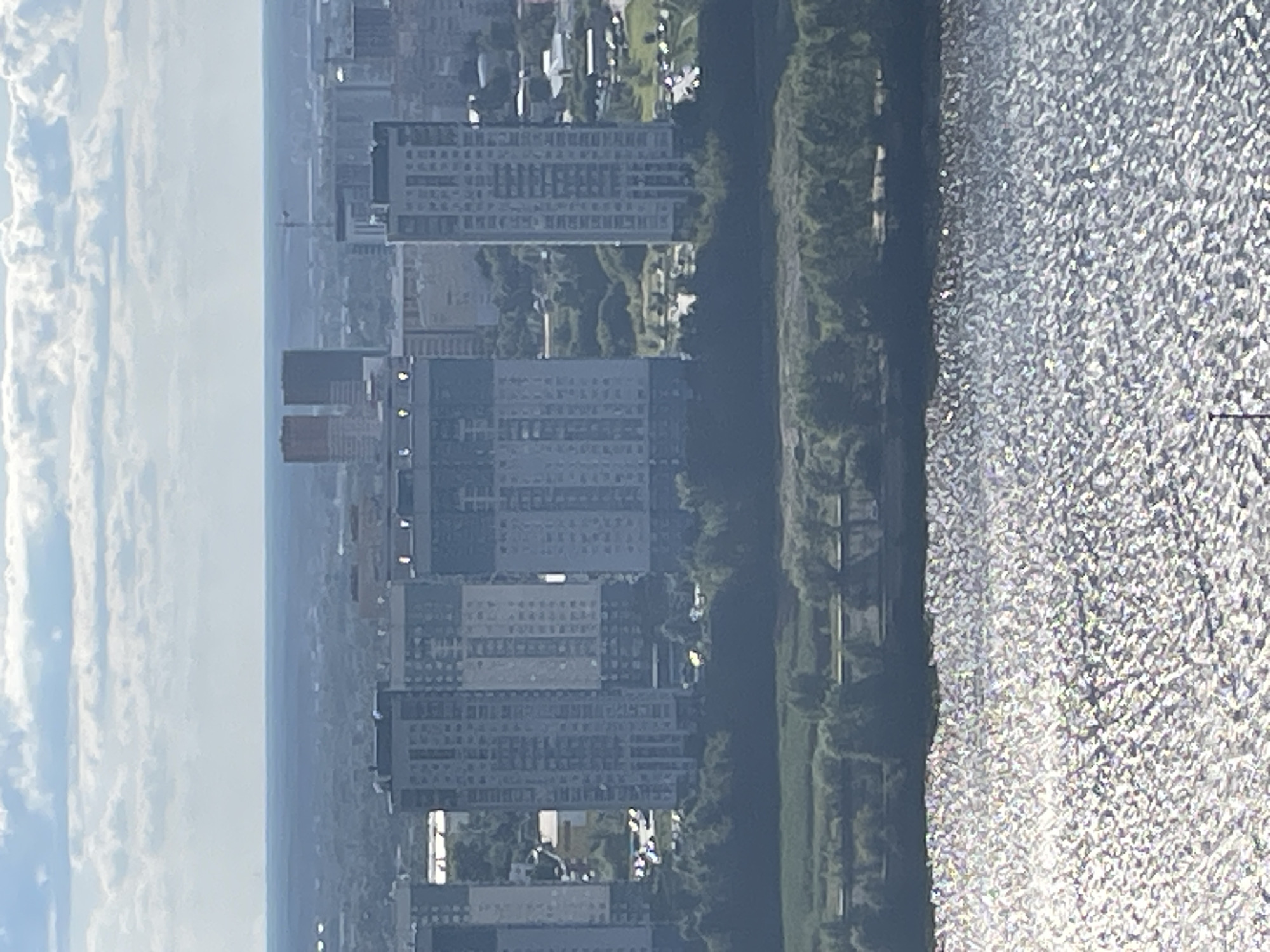The image shows a panoramic view of a city skyline with a body of water in the foreground. The water is highly reflective, with sunlight glinting off its surface. In the midground, there is a strip of lush green trees and vegetation along the bank. Beyond the trees, a series of modern, tall apartment buildings dominate the skyline. The buildings are characterized by their rectangular shapes and numerous windows, with some appearing to be light-colored and others darker. Further in the distance, a hazy cityscape stretches to the horizon, with more buildings and a faint outline of distant terrain under a sky filled with scattered clouds. The scene appears to be captured from a high vantage point, possibly from an aircraft or a very tall structure. The context suggests this is an urban area, likely residential given the presence of apartment complexes. Specifically, this image is a view of the newly developed area along the Oka River embankment in Nizhny Novgorod, Russia, often referred to as the "Oka-Volga Panorama". The buildings visible are part of the large-scale urban development projects on the left bank of the Oka River.