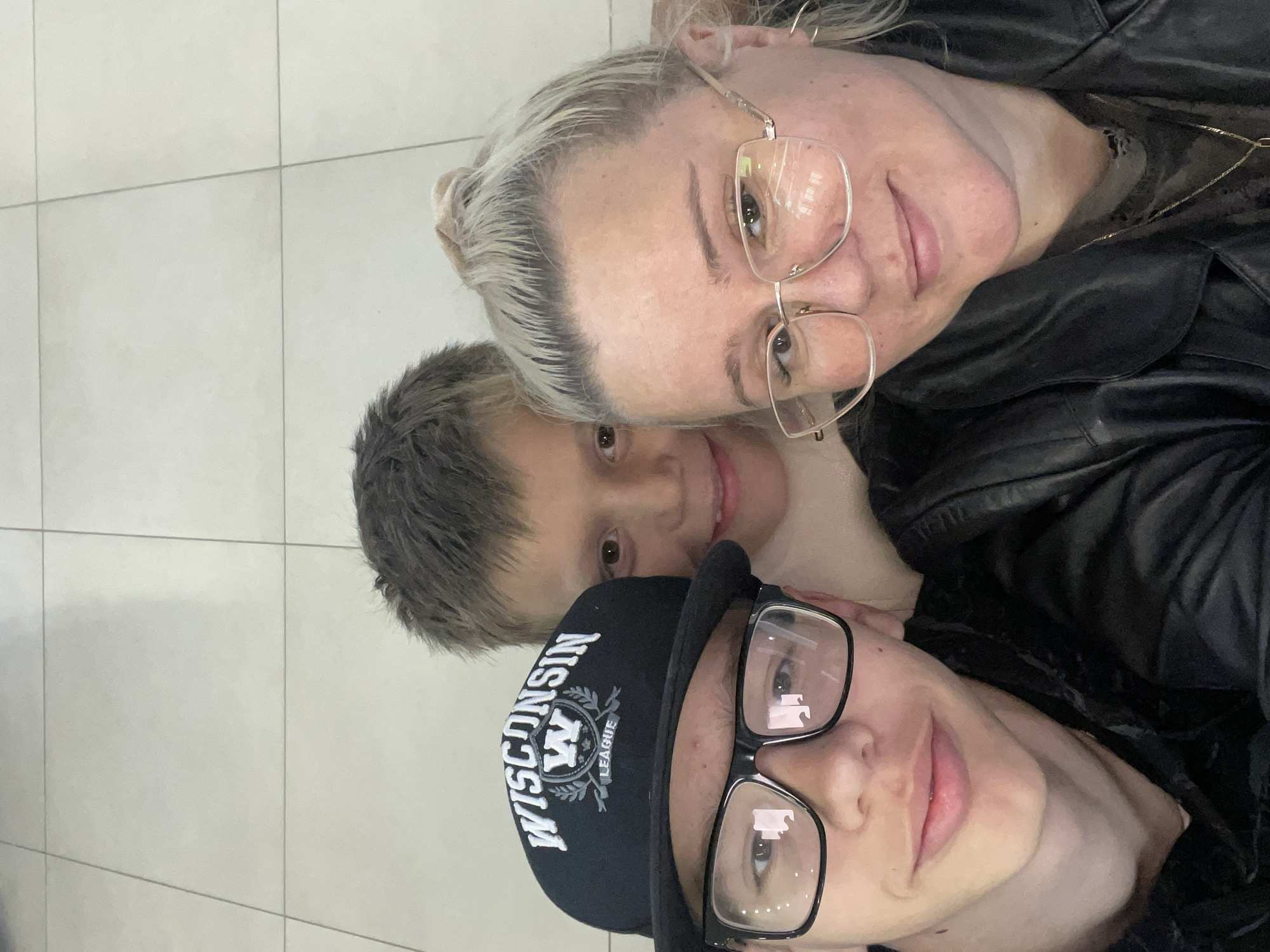 A selfie shows three people in a close-up shot, suggesting an indoor environment with tiled flooring. The person in the foreground left is a young male wearing glasses and a black baseball cap with "WISCONSIN LEAGUE" text. Next to him, a woman with blonde hair tied up and wearing glasses, and a black leather jacket is visible on the right. Behind them, a young boy with short brown hair is partially visible, smiling. The lighting appears to be bright, possibly from overhead sources. No specific location in Yekaterinburg, Russia is identifiable from the image.
