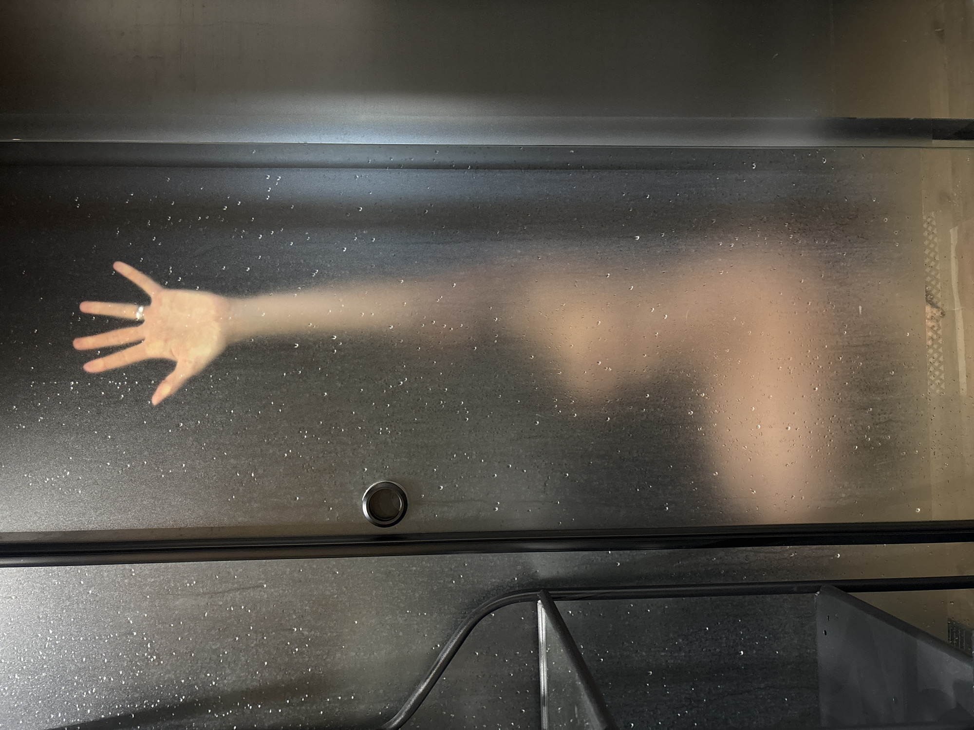 A hand with a ring is pressed against a translucent surface covered in droplets, presumably glass or plastic. The vague outline of a body extends from the hand. The environment appears to be a damp or humid enclosed space, possibly a shower or bathroom. The scene is metallic with droplets of water, and contains a round button and a ledge with a bar going across. There is a partial reflection of objects below. There is no information available to establish the location to be Chaikovsky, Russia.