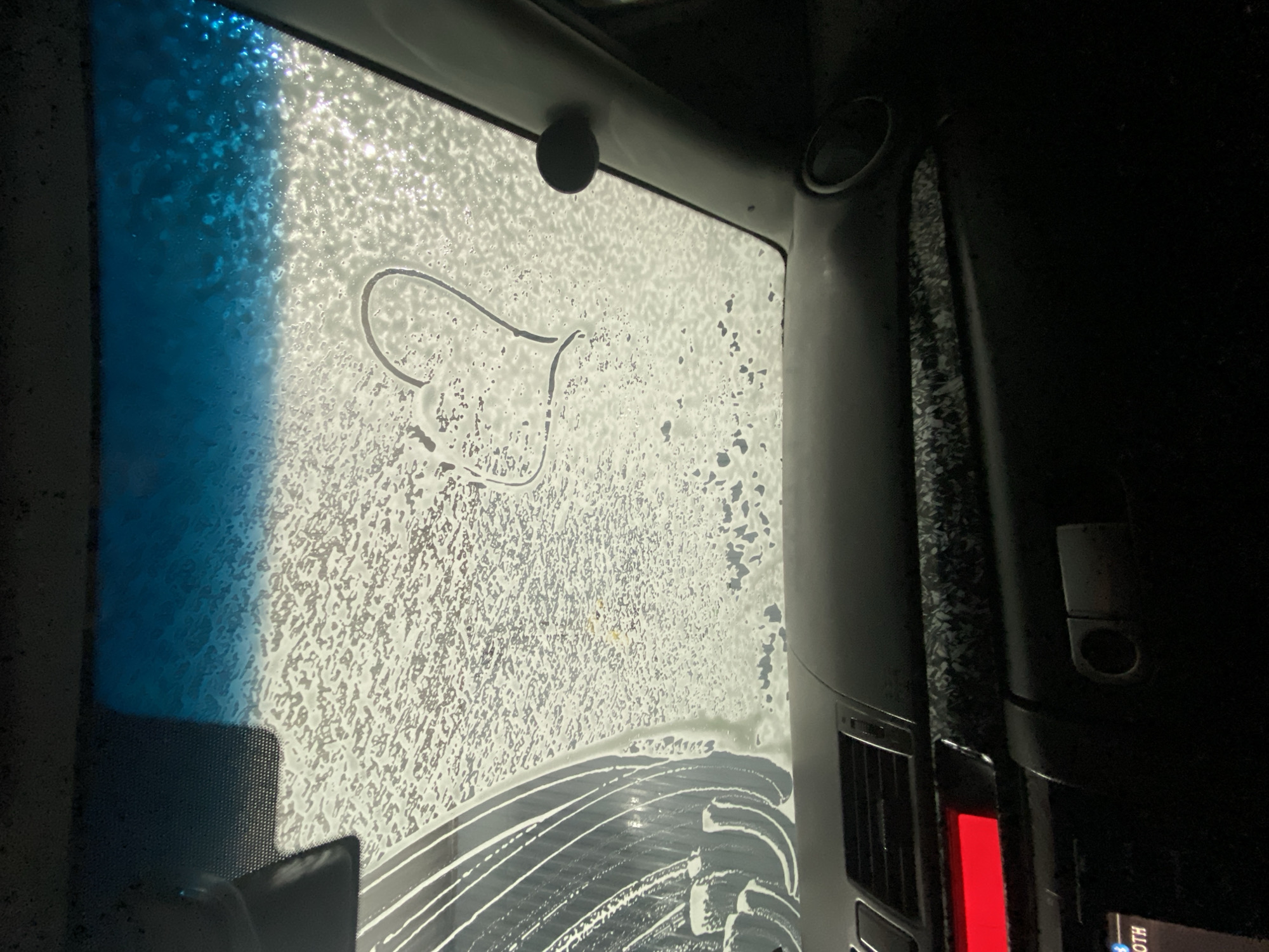 A view from inside a car during a car wash. The windshield is covered in soap suds, with a drawn heart visible. Part of the car's interior, including a vent and possibly the dashboard, is visible on the right. The outside light is visible through the soapy window. The location is not able to be determined from the image.