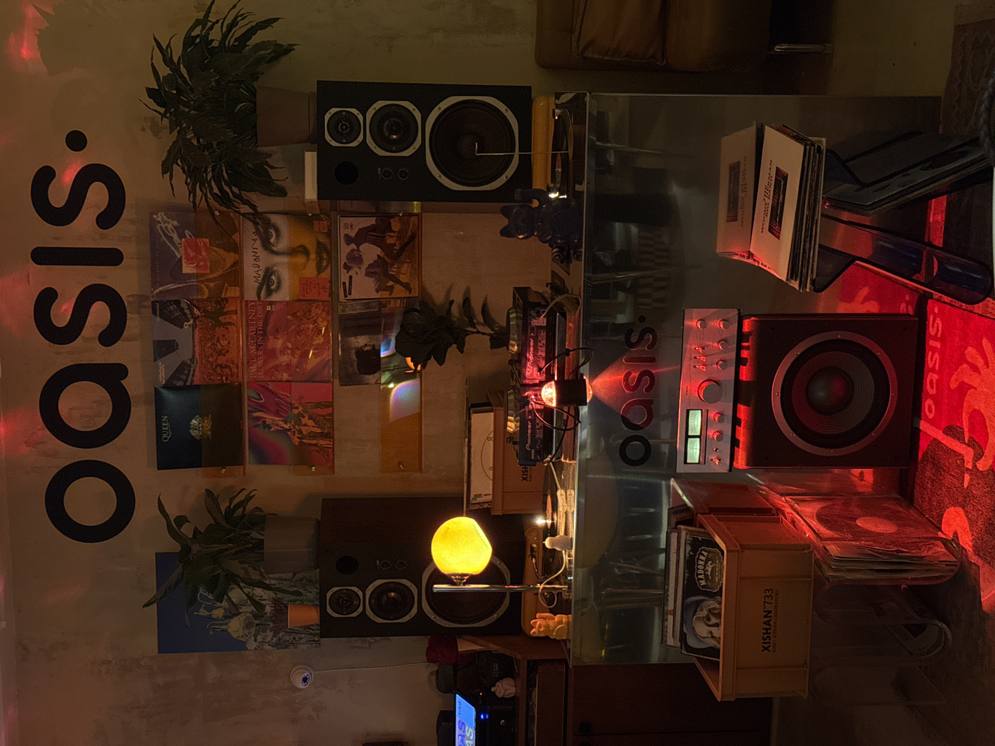 This image captures a cozy, warmly lit interior, likely a personal living room or dedicated music space in Krasnoyarsk, Russia, taken during the evening or night. The room is thoughtfully arranged for music appreciation, featuring a prominent audio setup and an extensive collection of vinyl records.
At the heart of the scene are two substantial, dark-colored speakers with multiple drivers, one on the left and another on the right. Between them, on a sleek, possibly glass surface, sits a silver Pioneer amplifier/receiver and other audio components. A striking spherical yellow lamp rests on the left speaker, providing a soft, localized glow. Additional ambient lighting comes from various red and white lights emanating from the audio equipment and surrounding decorations, creating an inviting atmosphere.
Numerous vinyl records are displayed across the room. Several album covers are clearly visible, including "MADONNA" (with "Madame X" and "Ray of Light" recognizable), "PAUL MCCARTNEY FLOWERS IN THE DIRT," and a "QUEEN" album. A wooden crate, filled with records, is inscribed with "XISHAN 733" and "©2011 XISHAN LABORATORY." Large, vertical "OASIS." text is a dominant feature on the left wall, with smaller "OASIS" branding repeated elsewhere. Potted plants are strategically placed, adding natural elements to the space. A small black cat-like figurine and other decorative items contribute to the personalized decor. The overall scene suggests a private sanctuary for music lovers, designed for relaxation and enjoyment. No individuals are present in the image.