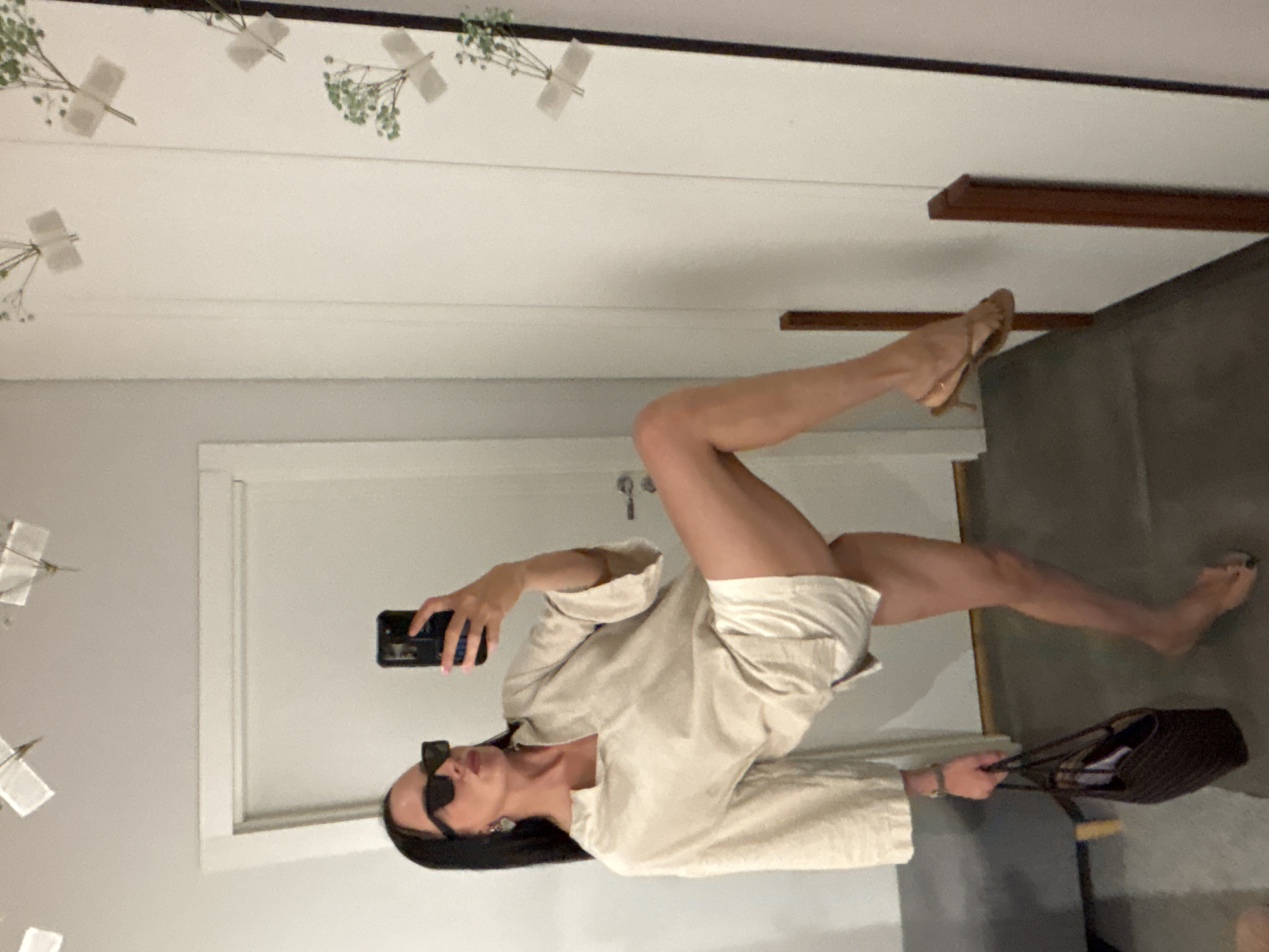 A person is taking a selfie in what appears to be an interior space, possibly a home. The person is wearing a light-colored shirt and shorts, sunglasses, and earrings. They are holding a smartphone in one hand and a brown purse in the other. One leg is bare and the other leg is in a heeled sandal. The background includes a white wall with taped flowers, a door, and a glimpse of a dark floor. The location is not discernible from this image.