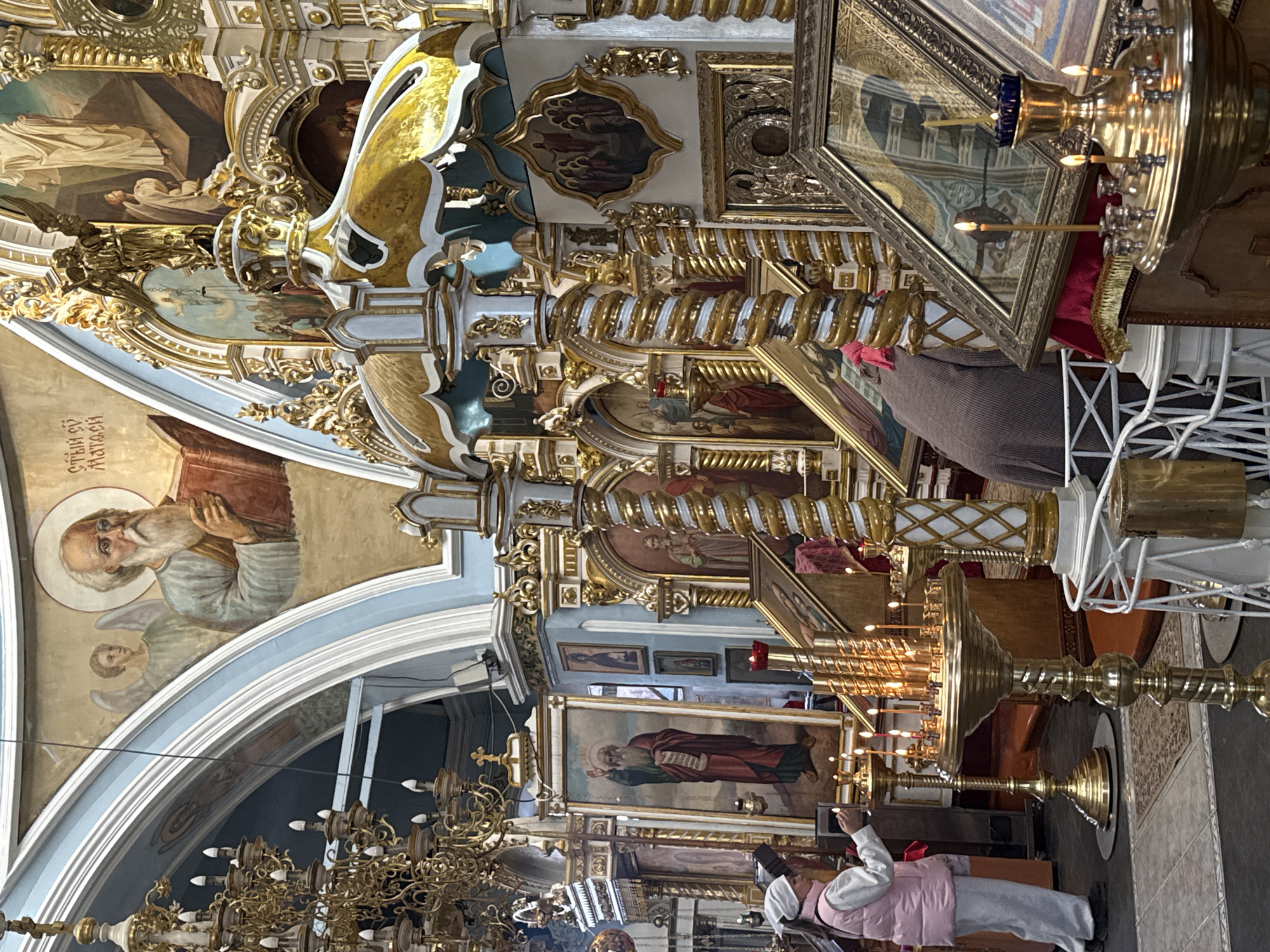 The image captures the interior of a Russian Orthodox church, likely located in Votkinsk, Russia. The architecture is ornate, with elaborate gold and white decorations, paintings, and chandeliers. A large painting of a bearded saint with a halo is visible on the upper left. Below, a woman in a white headscarf and pink vest is lighting candles on a golden stand. Other religious paintings and objects fill the space.