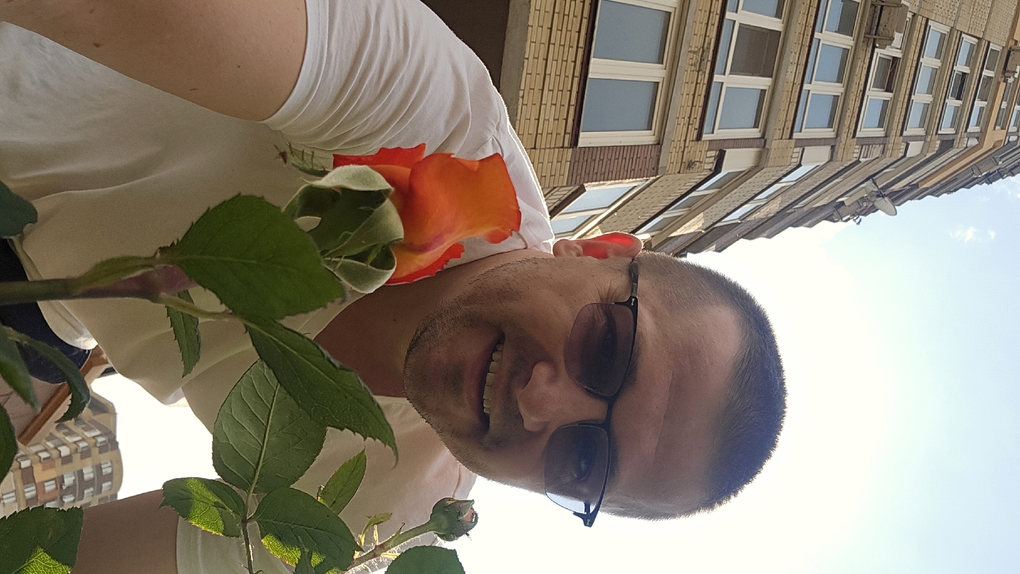 The image depicts a man in a white t-shirt wearing sunglasses, holding a bright orange rose in front of him. He is smiling at the camera. In the background, a tall, multi-story building with a brick facade and numerous windows is visible. The sky above is bright and appears to be daytime. The scene suggests an outdoor setting, possibly a balcony or yard, with a city view, consistent with a location in Kyiv, Ukraine.
