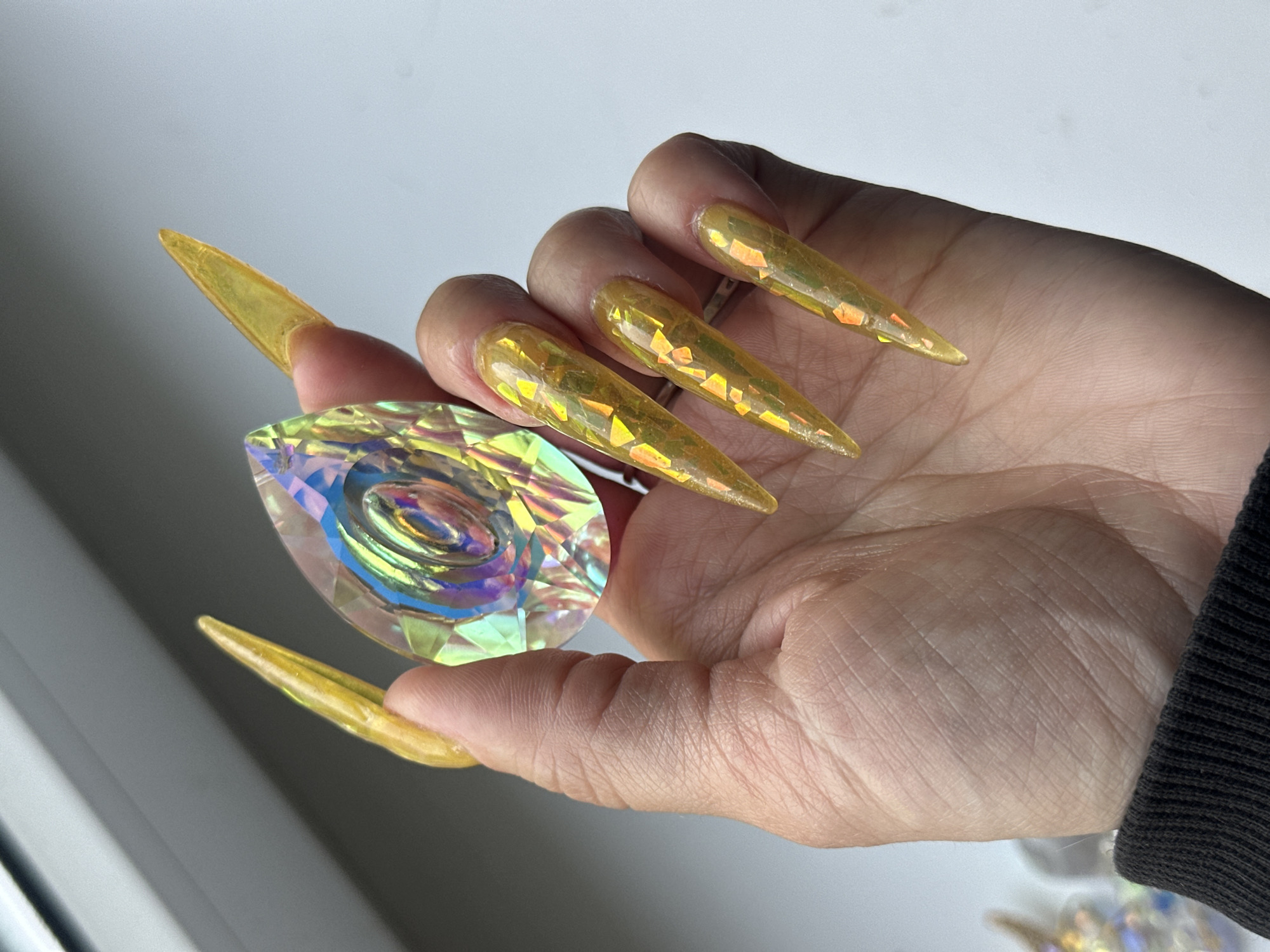 A human hand with long, yellow, stiletto-shaped fingernails is holding a multifaceted, teardrop-shaped crystal. The fingernails have iridescent flakes embedded within them. The hand is light-skinned with visible skin texture. The crystal displays various colors including blue, green, yellow, and purple. The background appears to be a white or light gray surface, possibly near a window. A black sleeve is visible at the lower right. The location and city are unknown.