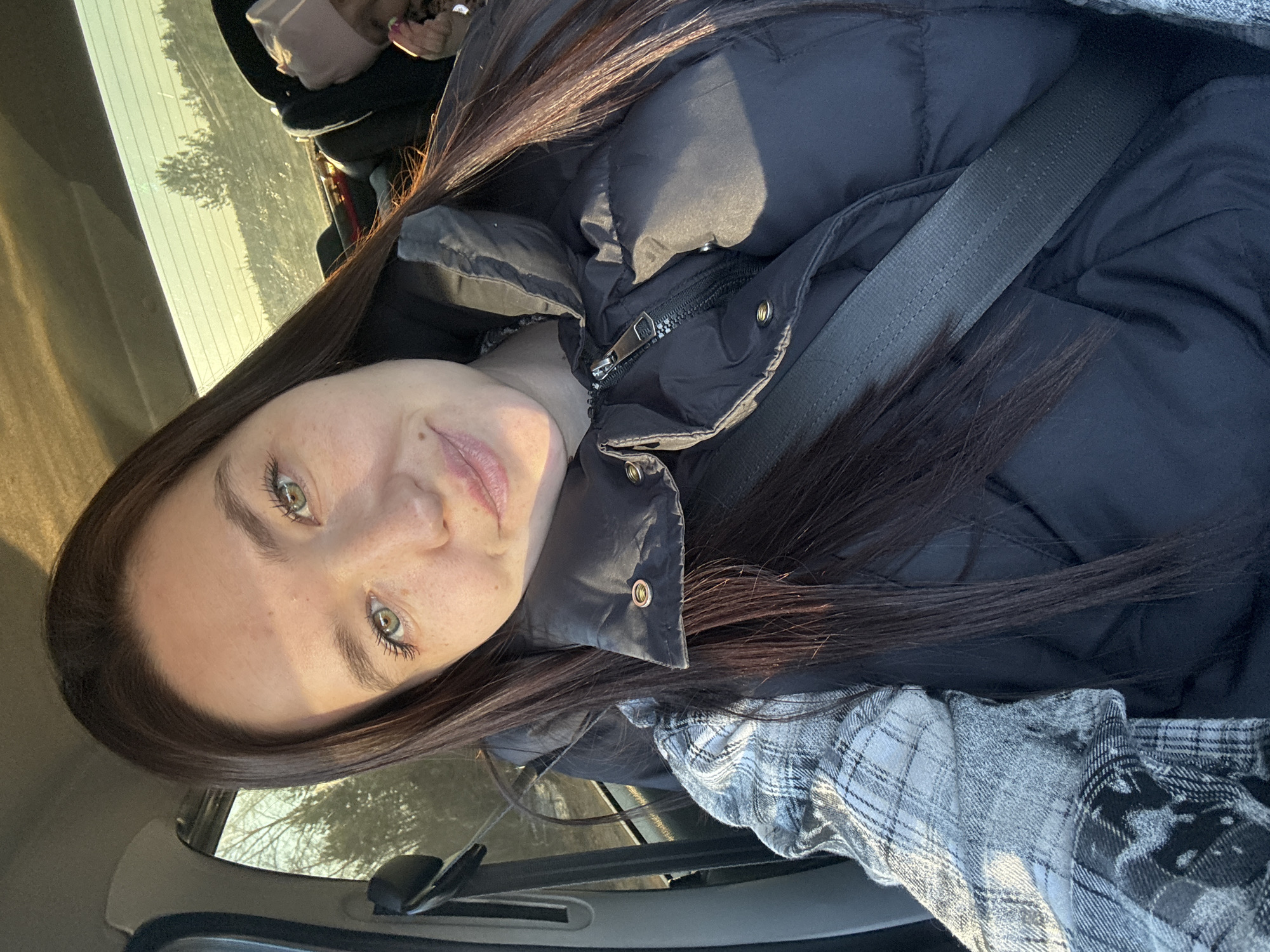 A selfie of a light-skinned young woman with long brown hair and green eyes is in a car, wearing a black puffer jacket and a gray plaid shirt underneath, secured by a seatbelt. The woman is looking directly at the camera with a neutral expression. Light is shining on her face. Part of the interior of the car is visible, including the dashboard and window with a view of trees and sky outside. There is no information to indicate that this image was taken in Гаревая, Russia.
