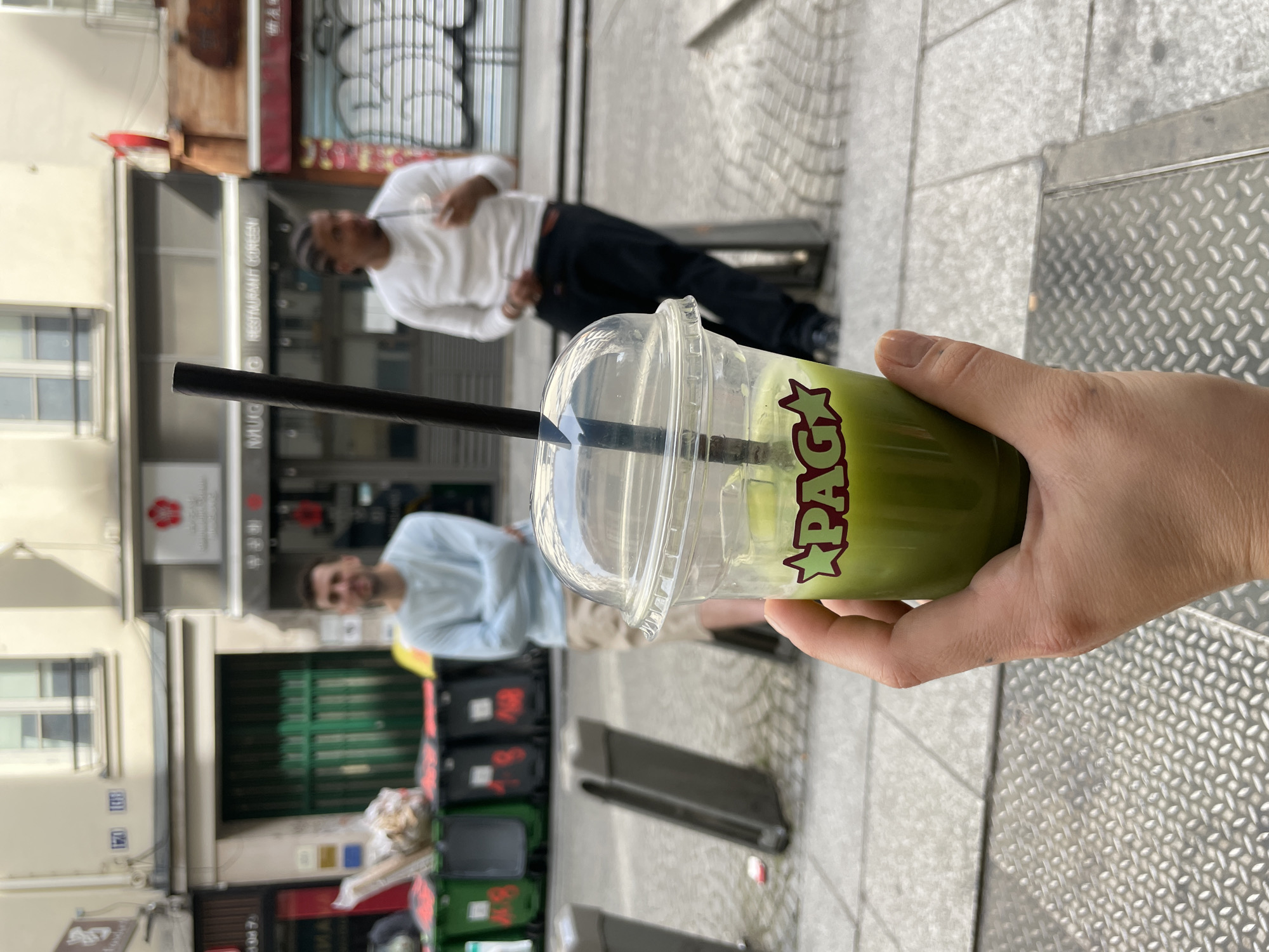 A person holds a plastic cup of green matcha-colored liquid with a black straw on a street in Paris, France. The cup has the word "PAGA" on the side. Two people are visible in the background; one is wearing a white shirt and dark pants and is sitting on a bench. The other person is wearing a light blue shirt and sitting on a bench. The sidewalk is concrete, and there are buildings and trash cans in the background.