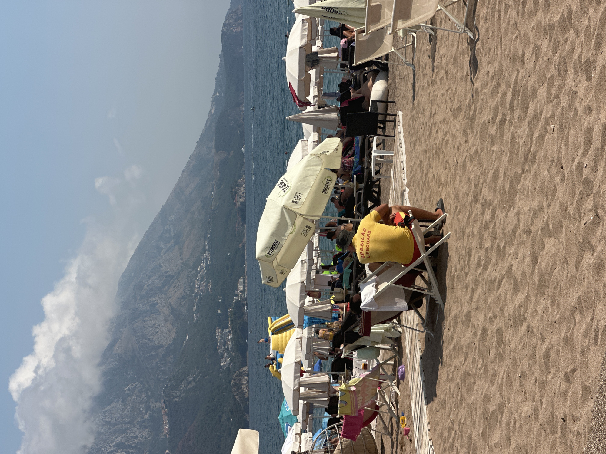 The image captures a bustling beach scene in Boreti, Montenegro, with a focus on the sandy shoreline populated by beachgoers. In the foreground, a man wearing a yellow shirt with "SPASILAC LIFESGUARD" printed on it sits on a chair, facing away from the camera, his legs crossed. The beach is lined with numerous white umbrellas providing shade for lounge chairs occupied by various individuals. Some people are visible relaxing on the chairs, while others are partially obscured. In the background, the azure sea stretches out, with a few boats visible in the distance. Beyond the water, a lush green mountain range rises, partially shrouded by clouds, with scattered houses and buildings dotting its slopes, indicating the presence of a town or village. A yellow inflatable water slide structure is also visible on the beach, with a few people on or around it. The overall atmosphere suggests a lively summer day at the beach.
