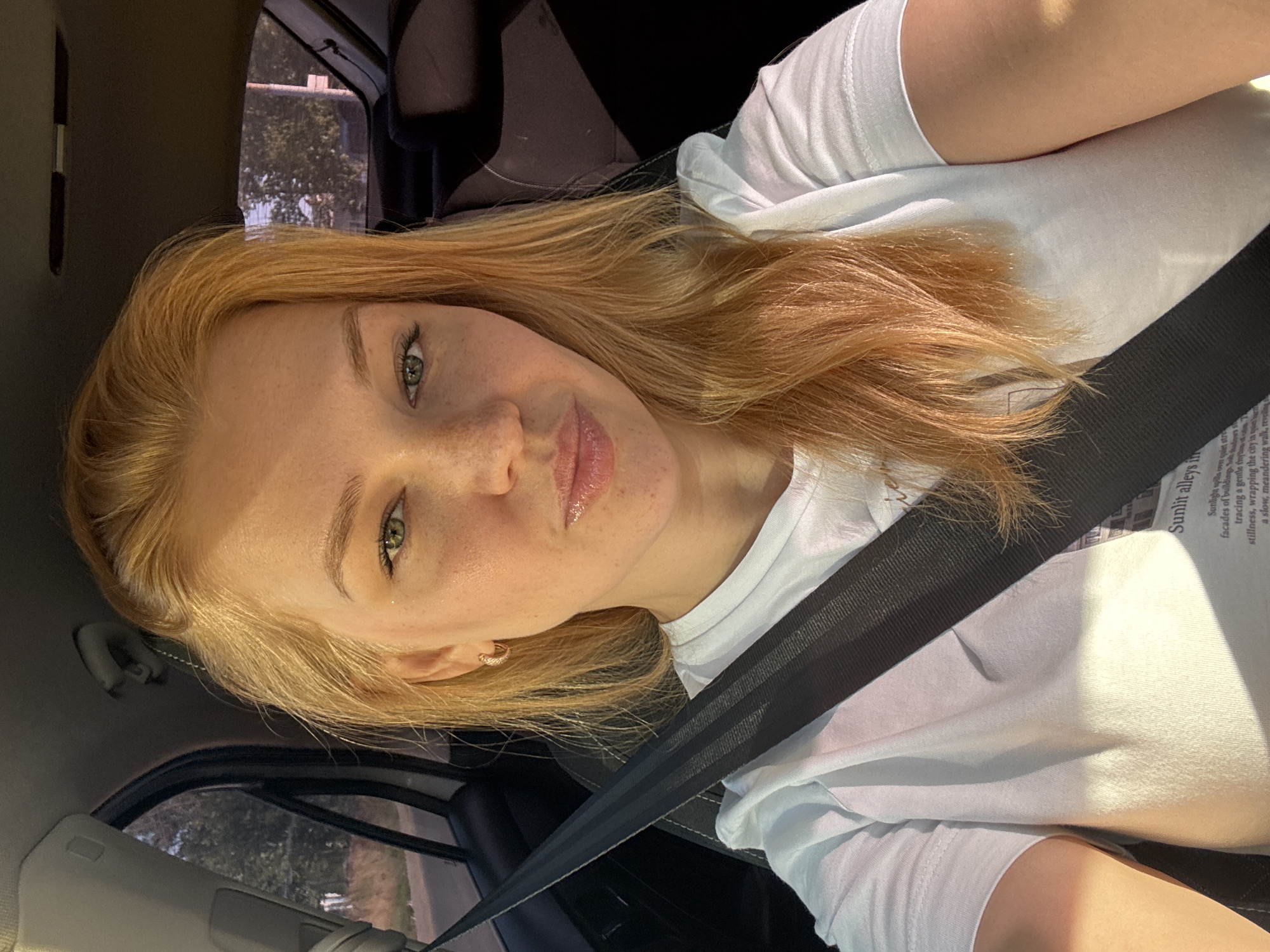 A young woman with blonde hair and green eyes, wearing a white t-shirt and a black seatbelt, is sitting in the passenger seat of a car. The car appears to be in motion, as indicated by the blurred background of trees and buildings. The woman is looking at the camera and smiling. The scene is set in the city of Zhukovo, Russia, during the daytime.