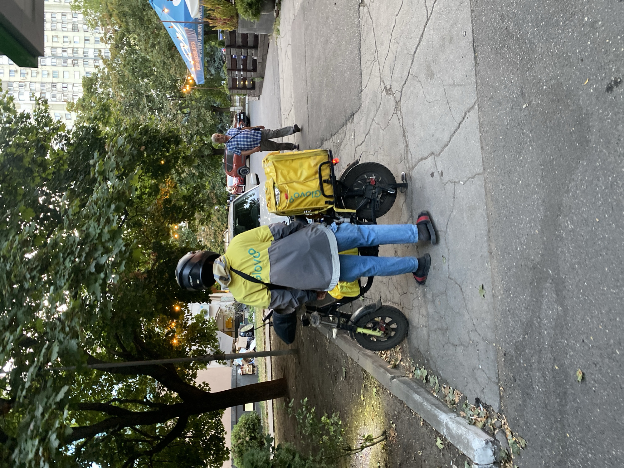 The image shows a delivery person standing next to their electric scooter, which is equipped with a large yellow insulated delivery bag labeled "Glovo". The delivery person is wearing a helmet, a yellow and gray jacket, blue jeans, and red and black slides. The scooter is parked on a cracked concrete sidewalk next to a street. In the background, there is a white car, a red SUV, a man in a blue checkered shirt, trees, buildings, and a blue awning. The scene appears to be in an urban environment, possibly a street or a parking area near a restaurant or cafe. The presence of the Glovo delivery bag and the electric scooter suggests a commercial delivery service is operating in this location within Chișinău, Moldova.