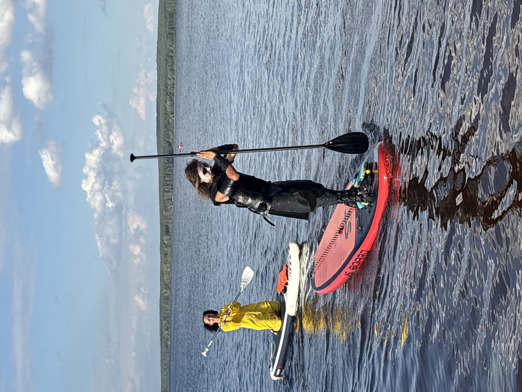 Two individuals are paddleboarding on a calm body of water. The person in the foreground, positioned on a red paddleboard, is facing away from the camera and leaning on a paddle. They have long, dark hair and are wearing dark, tactical-style clothing, including a vest and what appear to be knee-high boots. Their face is painted with white and black makeup.
The second person, on a white and grey paddleboard, is in the middle ground and facing the camera. They are kneeling and wearing a bright yellow, full-body suit. They are holding a paddle with a white and black blade.
The environment is a wide expanse of water with a forested shoreline visible in the distance. The sky is blue with scattered white clouds. The scene appears to be taking place on the Kama River near the city of Chaikovsky, Russia, as indicated by the presence of numerous oil and gas production facilities and pipelines in the background, which are characteristic of the industrial landscape in that region. The water is calm with ripples reflecting the sky and the paddleboarders.