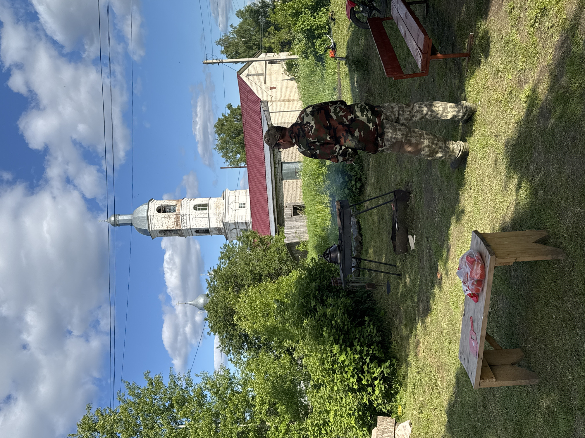 This outdoor image, captured on a sunny day in what appears to be a rural or village setting, depicts a man tending to a grill. The man is positioned slightly right of center, seen from behind and to his left, wearing camouflage trousers and a matching long-sleeved shirt with a cap. Smoke gently rises from a black metal grill or brazier in front of him, suggesting he is cooking.
The ground is covered in lush green grass, with patches of darker, possibly worn earth. To the man's right, a rustic wooden bench or table holds a clear plastic bag filled with red vegetables, possibly tomatoes or peppers, alongside a red spatula. Another empty wooden bench with red-painted legs is visible further back and to the right. Green trees and dense foliage form a natural backdrop across the scene.
In the background, a distinctive white bell tower of a church with a silver dome rises prominently on the left side of the frame. Another smaller, silver-domed structure is partially visible behind the trees further left. A building with a red roof and light-colored walls is situated behind the man, with more greenery and other structures in the distance. The sky is bright blue with scattered white cumulus clouds, and power lines stretch across the upper portion of the image. The overall atmosphere suggests a relaxed, summer day activity in a traditional Russian village setting, consistent with the provided location of Луптюг, Russia. No visible text is present.