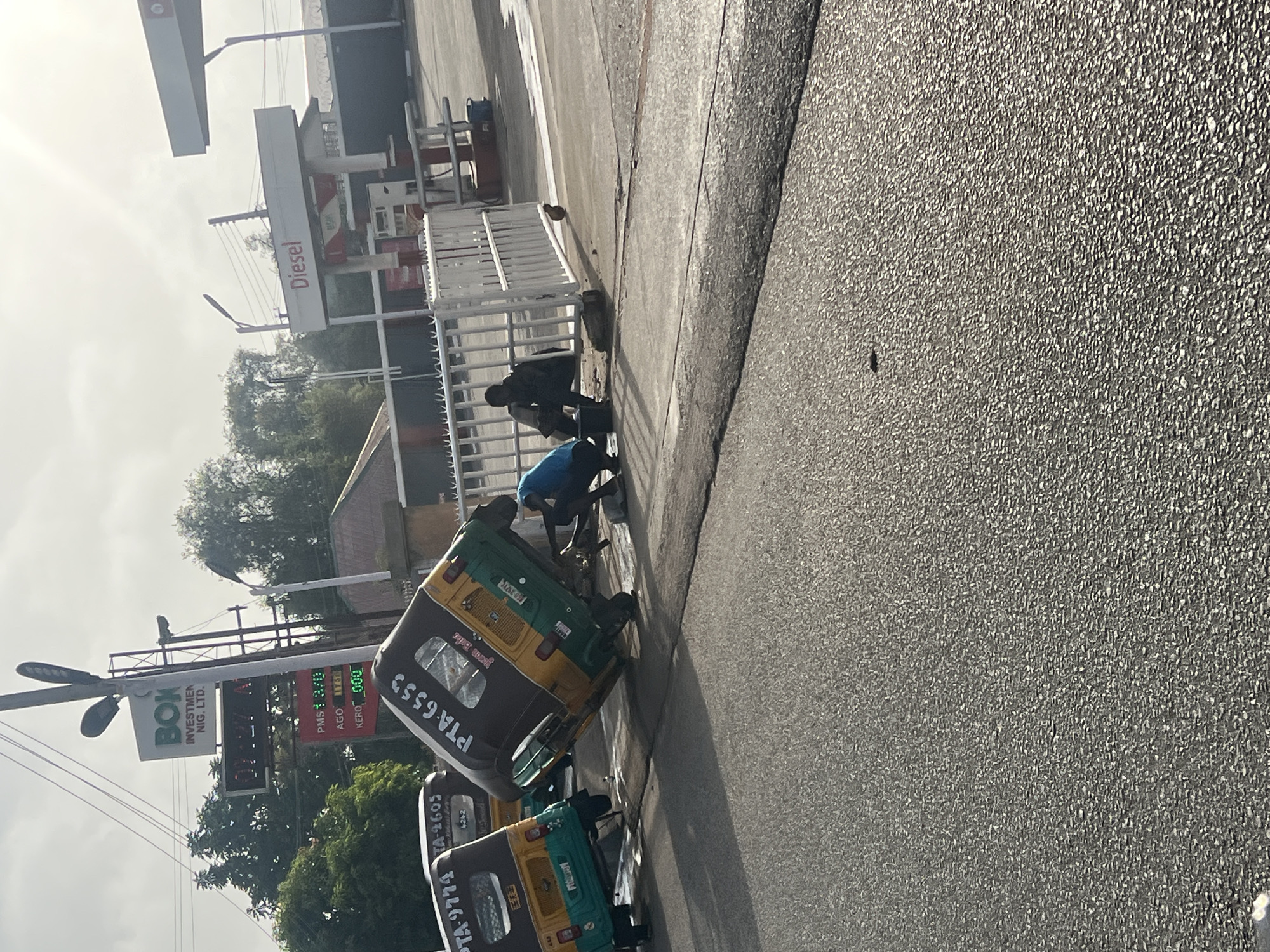 The image shows a street scene in Jos, Nigeria. Two auto-rickshaws (tuk-tuks) are visible, bearing license plates "PTA-9779" and "PTA-6553". The first reads TA-9779 and TA-2605 and 4242 below it. Next to the auto-rickshaws, two men are visible. One wears a blue shirt. The other wears a black shirt. They appear to be working near a white metal fence that separates the street from a gas station. The gas station has a sign that reads "Diesel". The gas prices are displayed. A sign that says "BOX INVESTMENT NIG. LTD" is visible above a sign with gas prices that read PMS188 AGO 13/0 KERO 000.