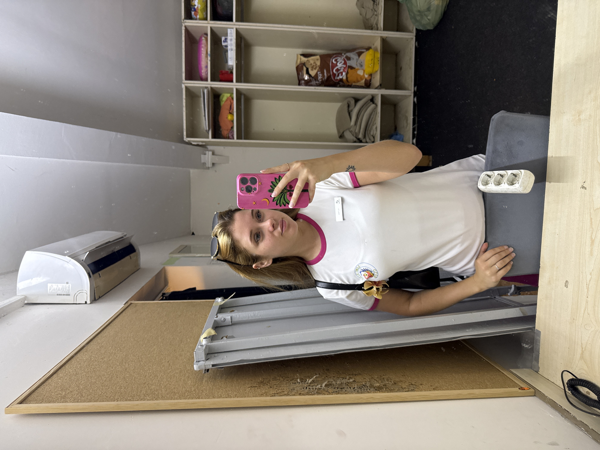 A young Caucasian woman with blonde hair, wearing a white polo shirt with pink trim, sunglasses on her head, and holding a pink smartphone with a palm tree design, is taking a selfie in what appears to be a locker room or backstage area. She has a name tag on her shirt and a flower pinned to her left shoulder. To her left, a corkboard is mounted on a wall, with a metal structure leaning against it. To her right, there are shelves filled with various items, including bags of snacks and folded fabrics. The floor is dark and the walls are light-colored. The presence of an air conditioning unit above the corkboard suggests an indoor, possibly climate-controlled, environment. The overall setting is utilitarian and functional.