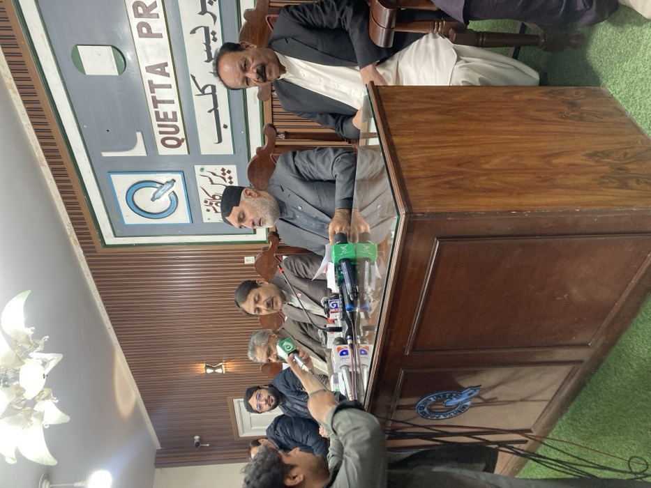 This image depicts an indoor press conference taking place at the Quetta Press Club in Quetta City Tehsil, Pakistan. Six men are visible within the frame.
The setting features a prominent wooden table with a glass top, behind which five men are seated in ornate wooden chairs. The wall behind them is clad in vertical wooden slats, with a large, framed sign displaying "QUETTA PRESS CLUB" in both English and Urdu, accompanied by a 'Q' logo. A multi-bulb chandelier hangs from the ceiling, and a wall sconce is visible on the left. The floor is covered with green artificial turf or carpet.
On the table, numerous microphones from various media organizations, including those labeled "Dawn News," "AP," "DM," "Express News," and one with a green logo, are positioned. Papers are also visible on the table surface. A 'Q' logo is affixed to the front panel of the wooden table.
In the foreground, a man with dark, curly hair, wearing a gray/green long-sleeved shirt, holds a microphone with a green logo towards the individuals seated behind the table. He appears to be a reporter or media personnel.
From left to right behind the table: a man in a dark jacket is partially visible; a man with a beard and glasses, wearing a dark jacket, is seated; a man with a mustache, dressed in a light brown suit jacket over a collared shirt, is actively speaking into the microphones; a man with a dark beard and a black cap (karakul style), wearing a gray suit over dark traditional attire, holds papers in his left hand; and a man with a prominent mustache, wearing a dark suit jacket over a white shalwar kameez, is seated. A sixth man is partially visible on the far right edge of the frame.