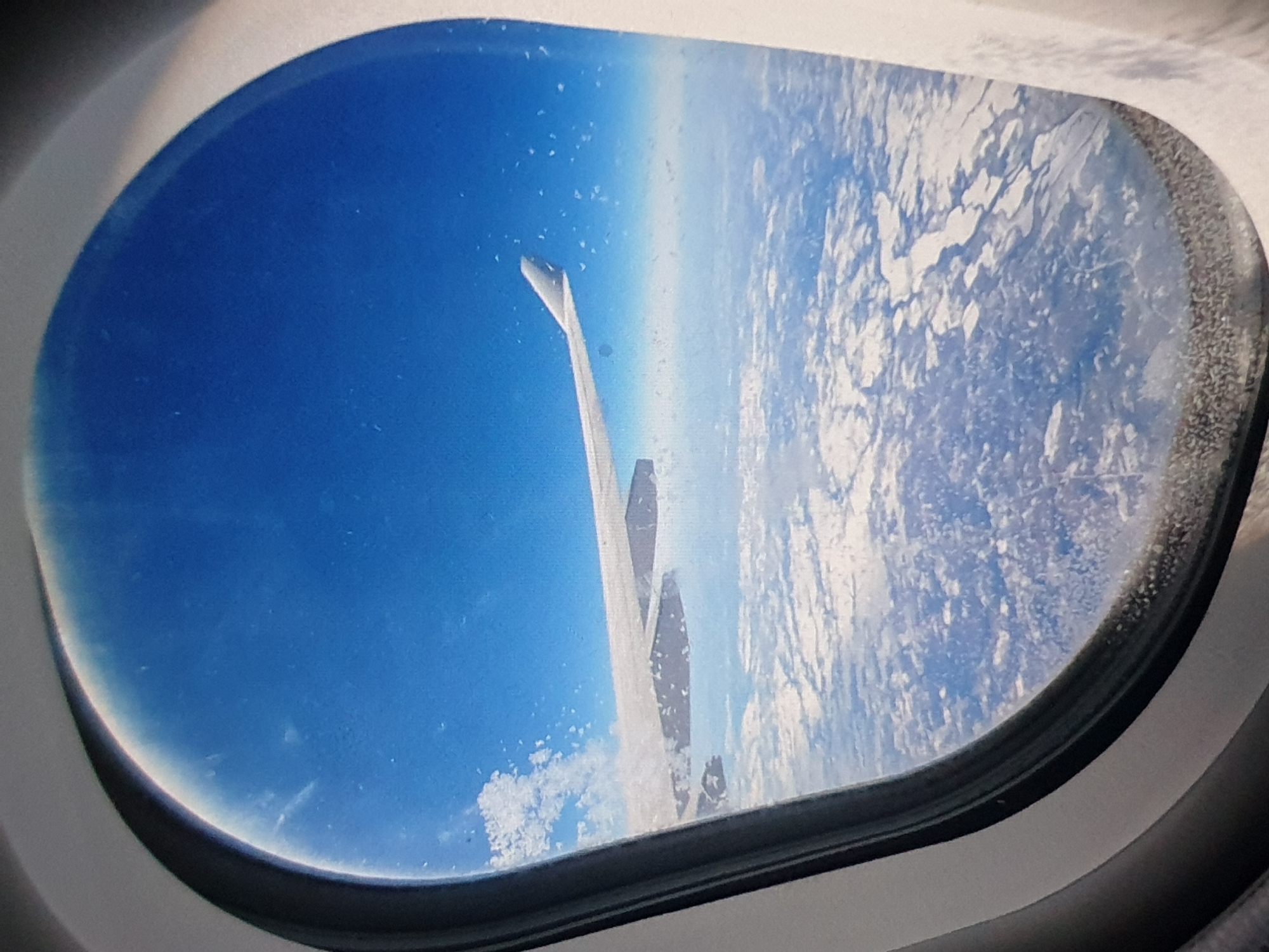 A view from an airplane window shows a wing and the Earth below. The sky is blue and clear, and a layer of clouds is visible. The location is Unknown, Unknown.