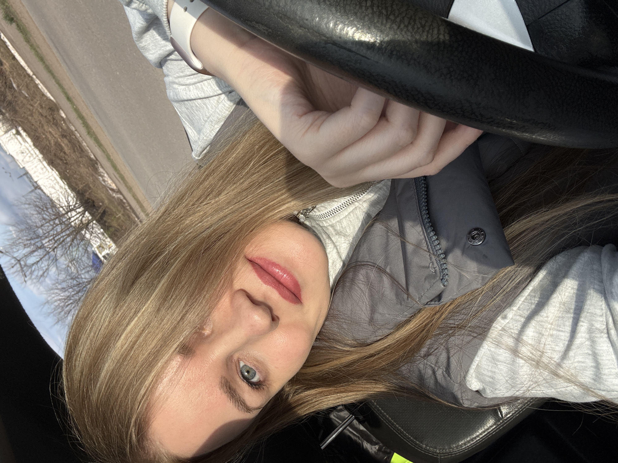 A woman with blonde hair and blue eyes is posing in what appears to be the driver's seat of a car, with her hands on the steering wheel. She is wearing a grey zip-up jacket over a lighter grey long-sleeved shirt. Outside the car window, a barren landscape with leafless trees and a road can be seen. The photo's geographical location is unspecified.