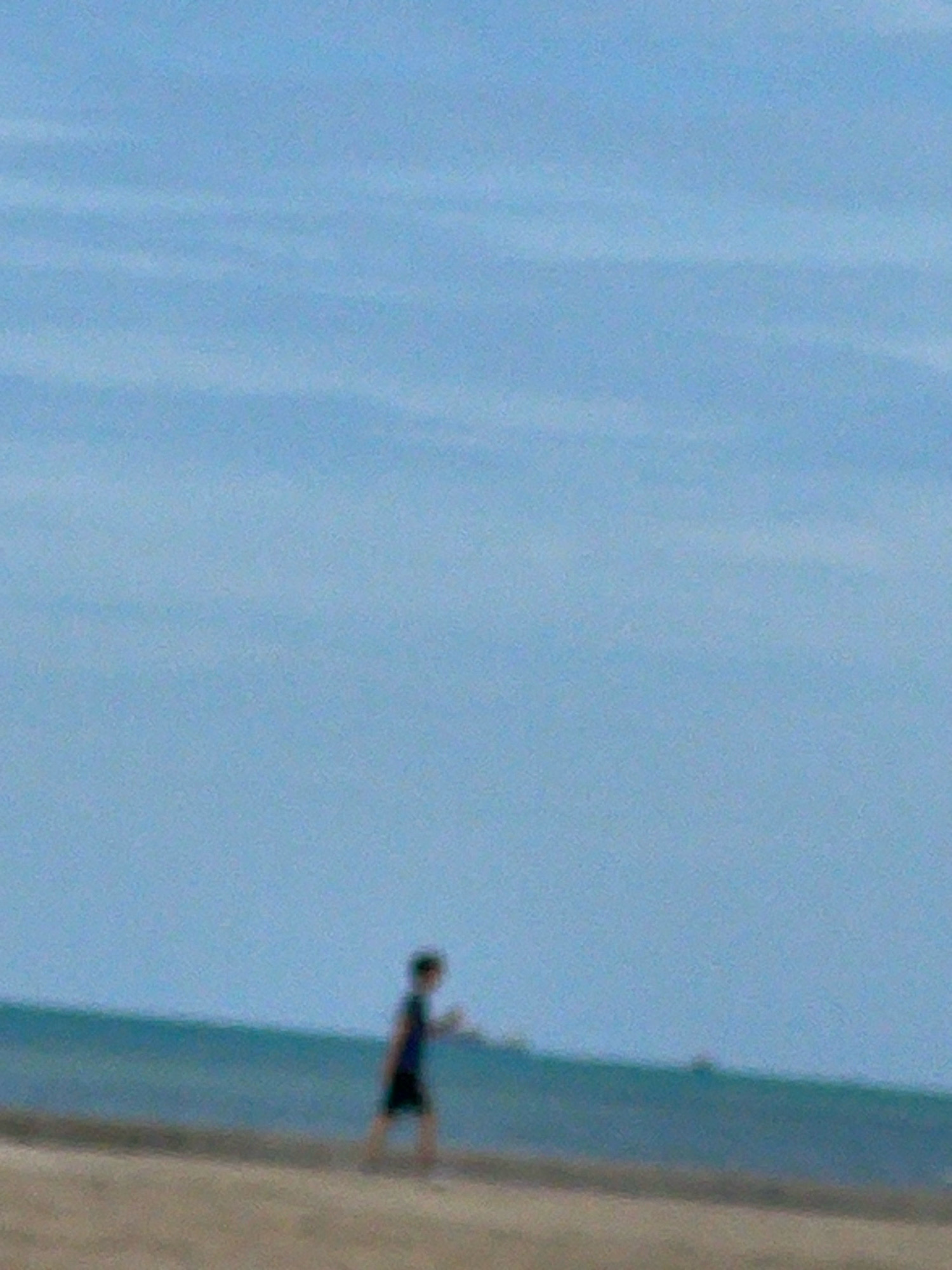 A lone person, appearing to be a child, walks on a sandy beach beside a calm ocean under a hazy blue sky. The individual is seen in silhouette, wearing dark shorts and a dark shirt. The person's posture suggests they are in motion, possibly walking towards the right of the frame. The background consists of the ocean, with a faint, blurry line of the horizon where the water meets the sky. The foreground is the beach sand. The overall scene is slightly out of focus and appears to be taken from a distance. This location is consistent with the coastal areas of Ban Na Chom Thian, Thailand, known for its beaches and ocean views.
