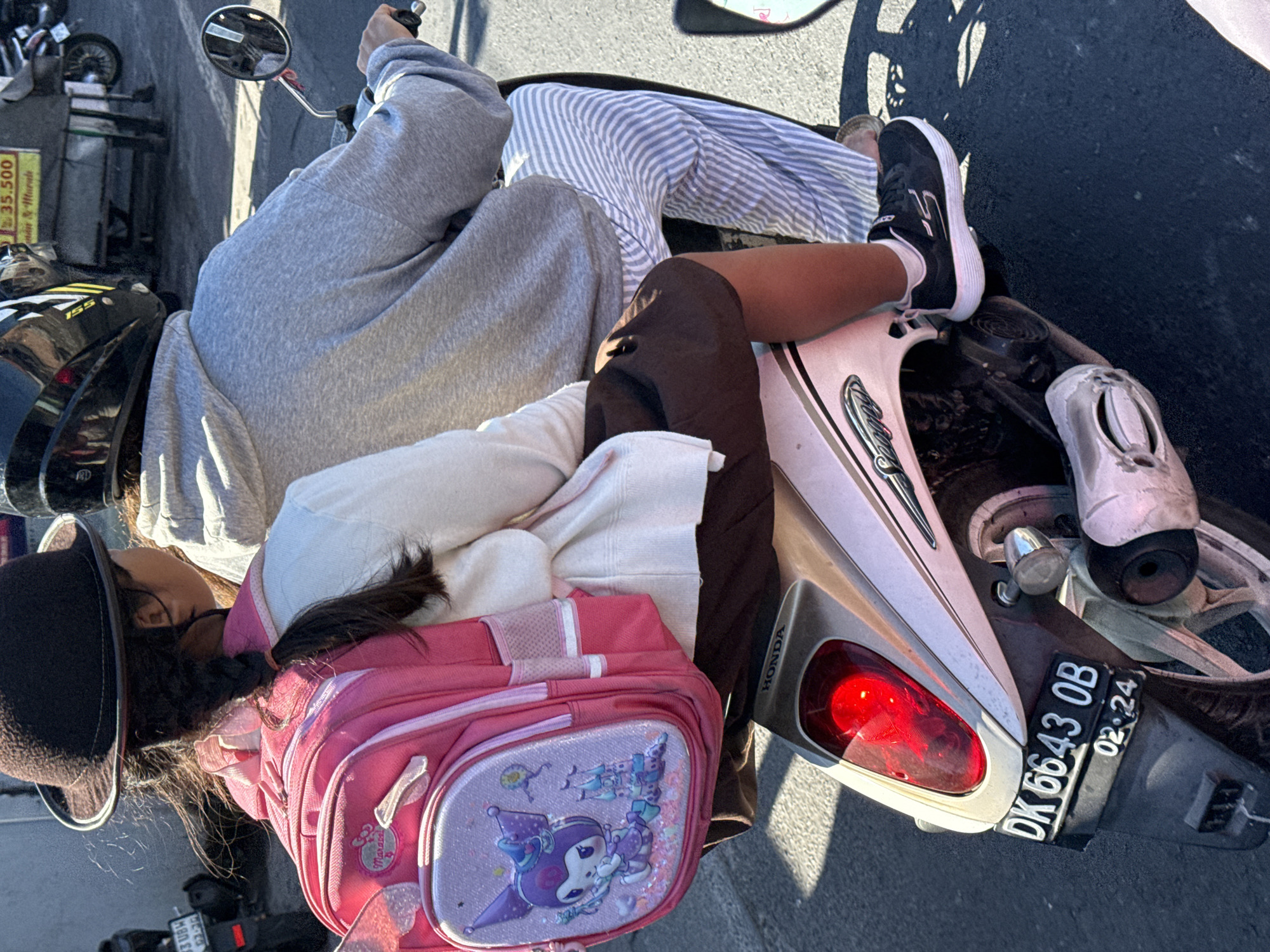 Two people are riding a motorcycle on a paved road in Denpasar, Indonesia. The person in front is a young girl wearing a brown hat, a white sweater, and a pink backpack with a cartoon character on it. She is looking to her right. The person behind her is wearing a grey hoodie and striped pants. Their hands are not visible, but the person behind appears to be holding onto the driver. The motorcycle is silver and has a red taillight. A license plate that reads "DK 6643 OB 02.24" is visible on the back of the motorcycle. The lighting suggests it is daytime.