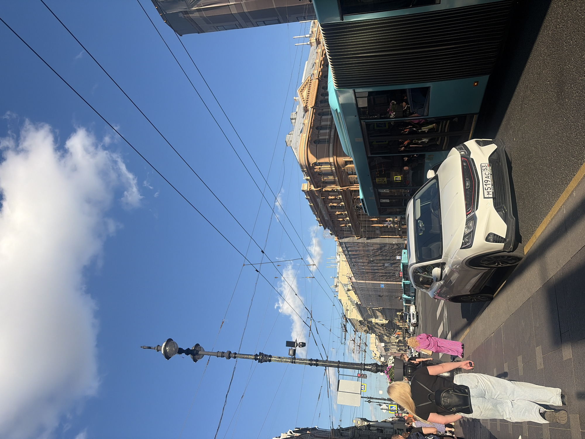 An eye-level, medium-shot image shows a street scene in Saint Petersburg, Russia, on a bright, sunny day. The sky is a vibrant blue with scattered white clouds. The perspective is from the sidewalk, looking down a street lined with buildings and a tram.
In the foreground, on the left, a woman with long blonde hair, wearing a black t-shirt and light-wash jeans, is walking away from the camera. She has a black backpack on her back. Further down the sidewalk, a person in a bright pink patterned outfit is walking towards the right. Other pedestrians are visible in the distance on the sidewalk.
Dominating the street is a modern, teal-colored articulated tram. In front of the tram, a white SUV with a license plate reading "M 519 AC 53" is stopped. The street is paved with asphalt, and a yellow line marks the edge of the lane.
The buildings lining the street are of various architectural styles and colors, with ornate detailing and multiple stories. Overhead, a complex network of tram and utility wires crisscrosses the sky, suspended from tall, dark lampposts. One lamppost, elaborately decorated, is prominently featured on the left side of the frame. The architecture and tram system suggest a historic urban setting. The overall impression is one of a bustling city street on a pleasant day.