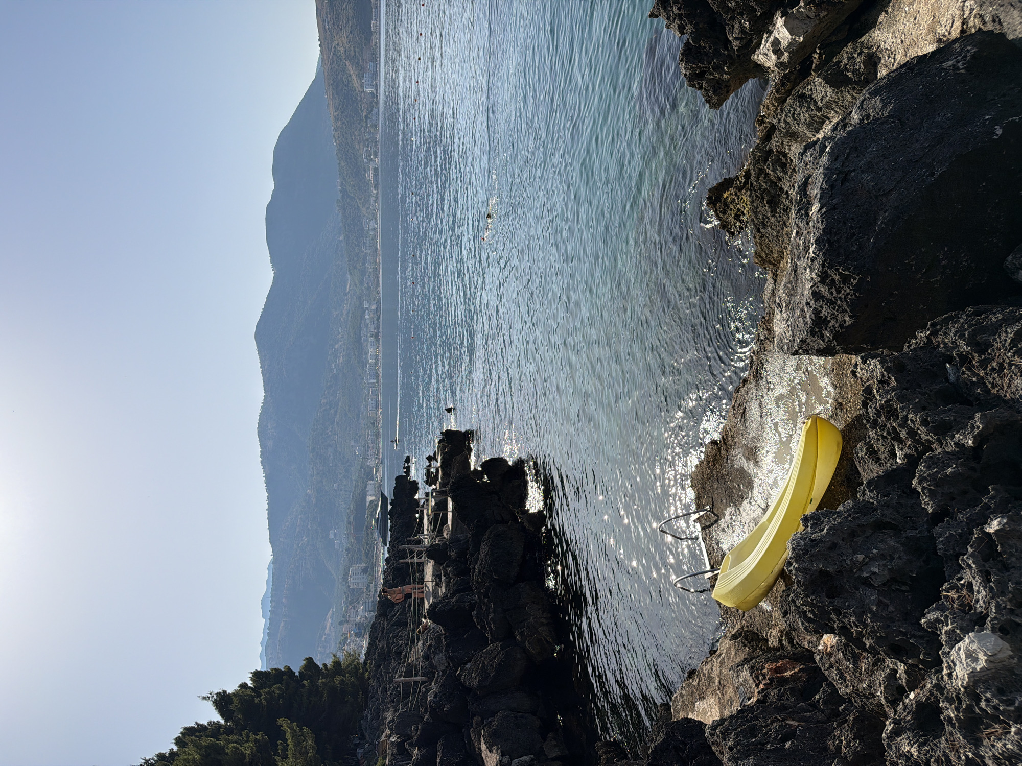 A bright, sunny day at a rocky coastline in Himarë, Albania. The foreground features dark, jagged rocks with a yellow kayak resting on the shore, partially submerged in the clear, turquoise water. Metal ladder rungs are visible in the water near the kayak. In the midground, a person stands on a wooden platform overlooking the sea, while another person appears to be swimming in the water. The background reveals a town with white buildings nestled against green, rolling hills under a clear blue sky. Several boats are visible on the water, and buoys mark a swimming area. The overall scene depicts a tranquil coastal setting with elements of leisure and natural beauty.