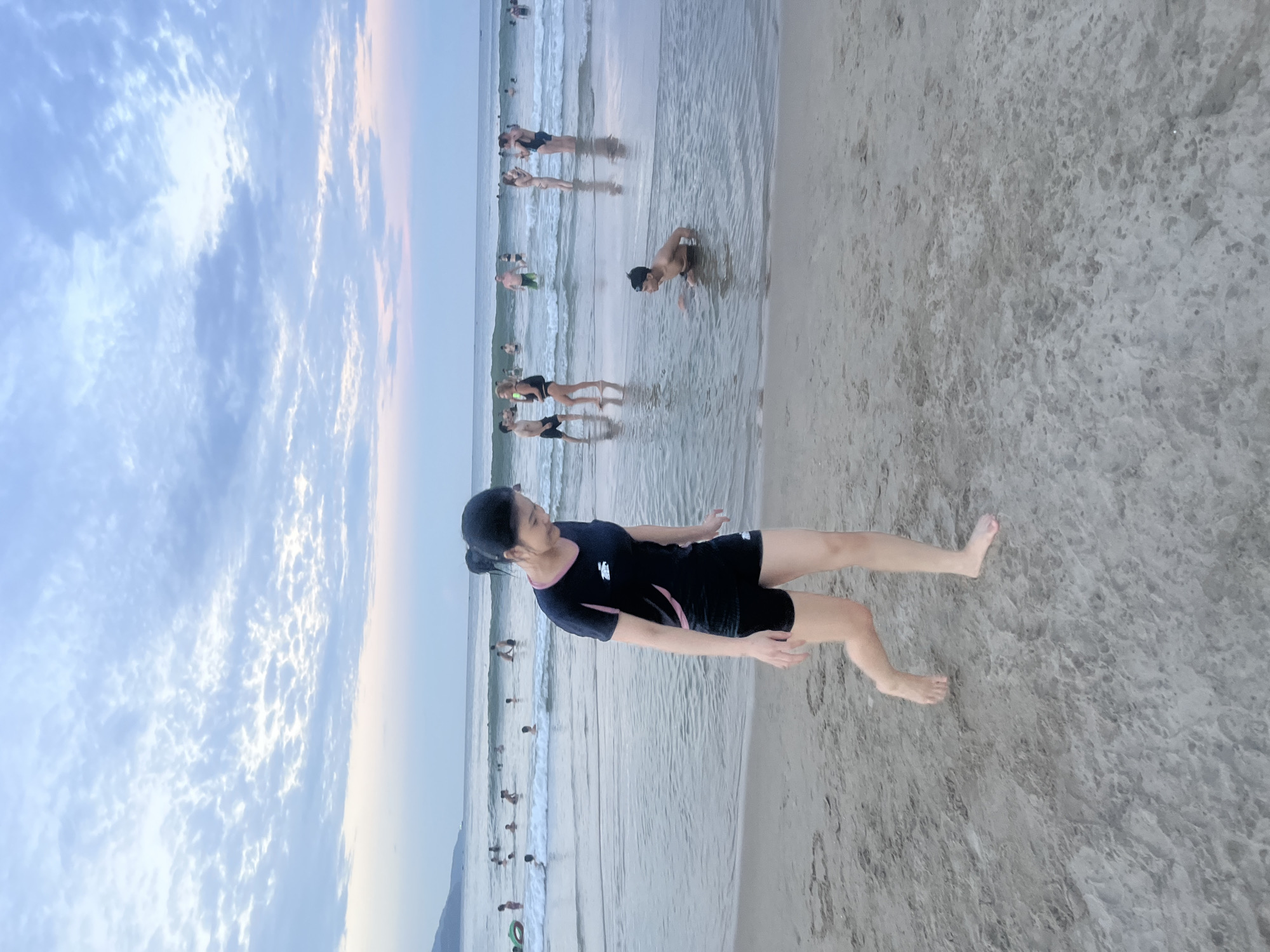 The image depicts a busy beach scene in Da Nang, Vietnam, with many people enjoying the water and the sand. In the foreground, a woman in a black and pink athletic swimsuit walks along the wet sand, her gaze directed downwards and to her right. Behind her, the ocean is populated with numerous individuals, some wading, some swimming, and some interacting with each other. A young man is visible in the middle ground, sitting in the shallow water. The sky above is filled with scattered clouds, suggesting either early morning or late afternoon light. The overall impression is one of a lively, recreational environment.