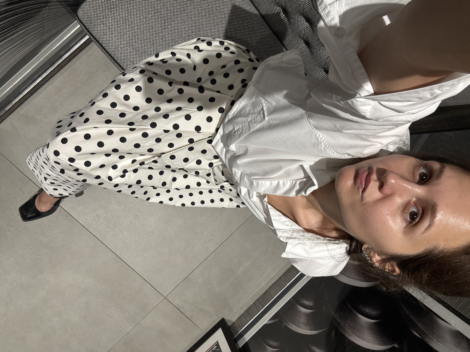 A woman is captured in a selfie, looking directly into the camera from an upward angle. She has medium brown hair, brown eyes, and a neutral expression, with her skin showing a slight sheen. She is wearing a crisp white button-down shirt and a flowing white skirt or wide-leg pants patterned with black polka dots. Her left foot, clad in a black flat shoe with a square toe, is visible towards the bottom left.
The setting is a modern indoor space, potentially an apartment or hotel room in Dubai, United Arab Emirates. The floor is composed of large, light gray tiles with visible grout lines. A dark gray upholstered chair or sofa with a subtle geometric pattern (possibly houndstooth) is seen behind and to her right. The background also features a dark, reflective surface, possibly a piece of furniture, and a partially visible framed artwork with abstract, dark circular motifs. A silver-colored trim is also visible near the floor. The lighting appears to be artificial or diffused natural light. No visible text is present.